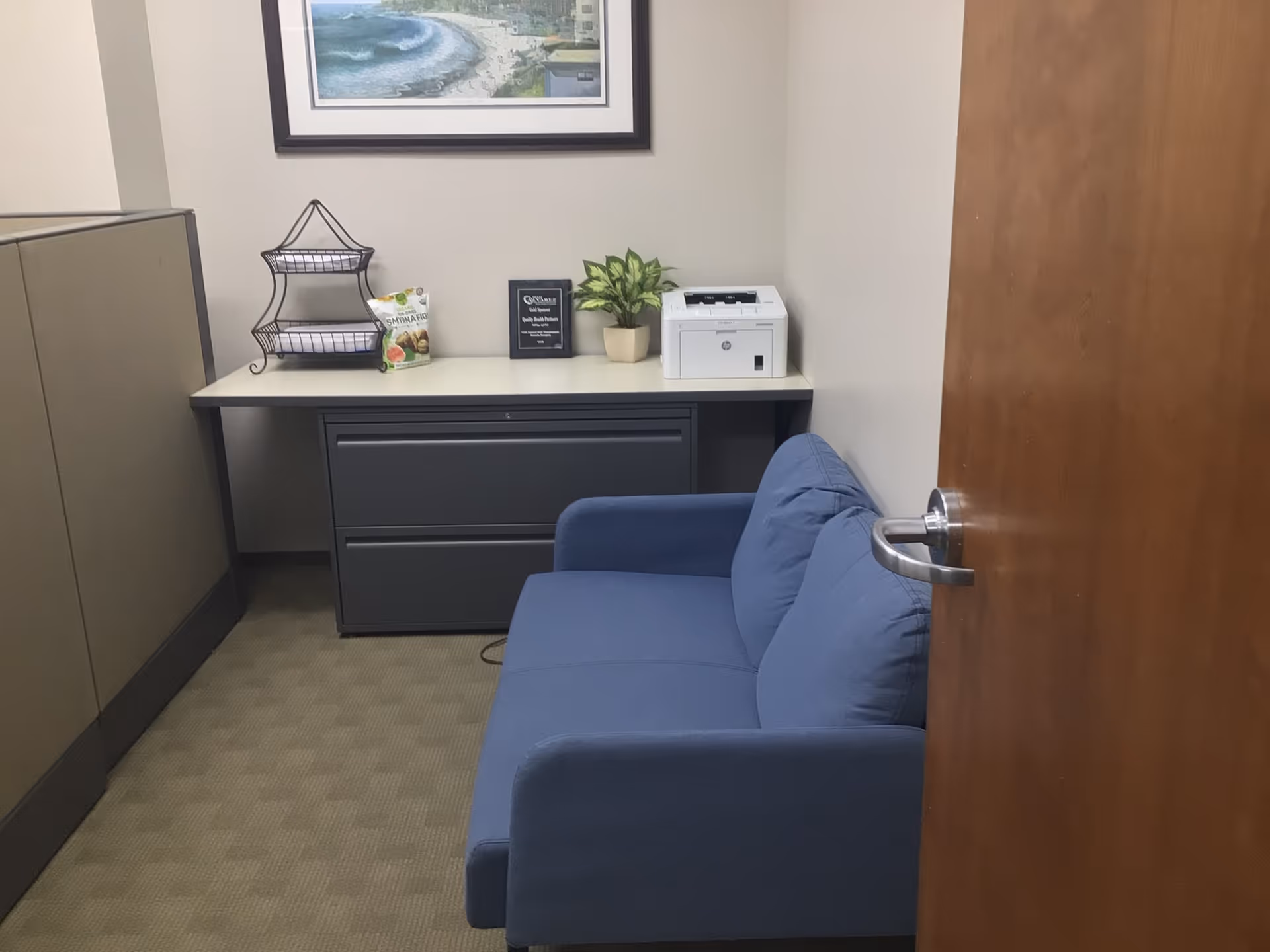 Interior office lounge with blue sofa, filing cabinet, printer, plant, and framed wall art.
