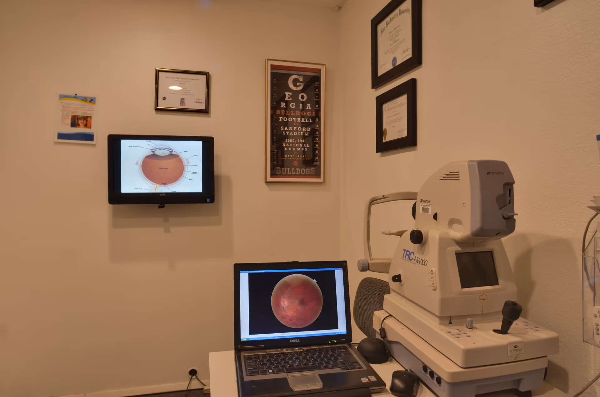 An optometry examination room with medical equipment, a laptop, and framed certificates on the wall.