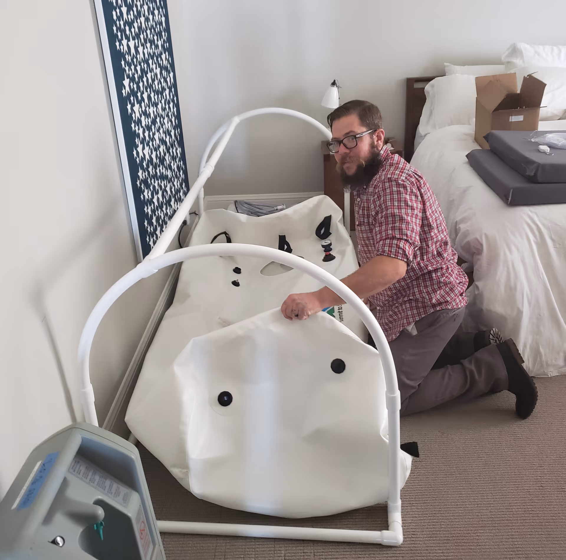 Man kneels beside a large white hyperbaric chamber in a bedroom with a bed.
