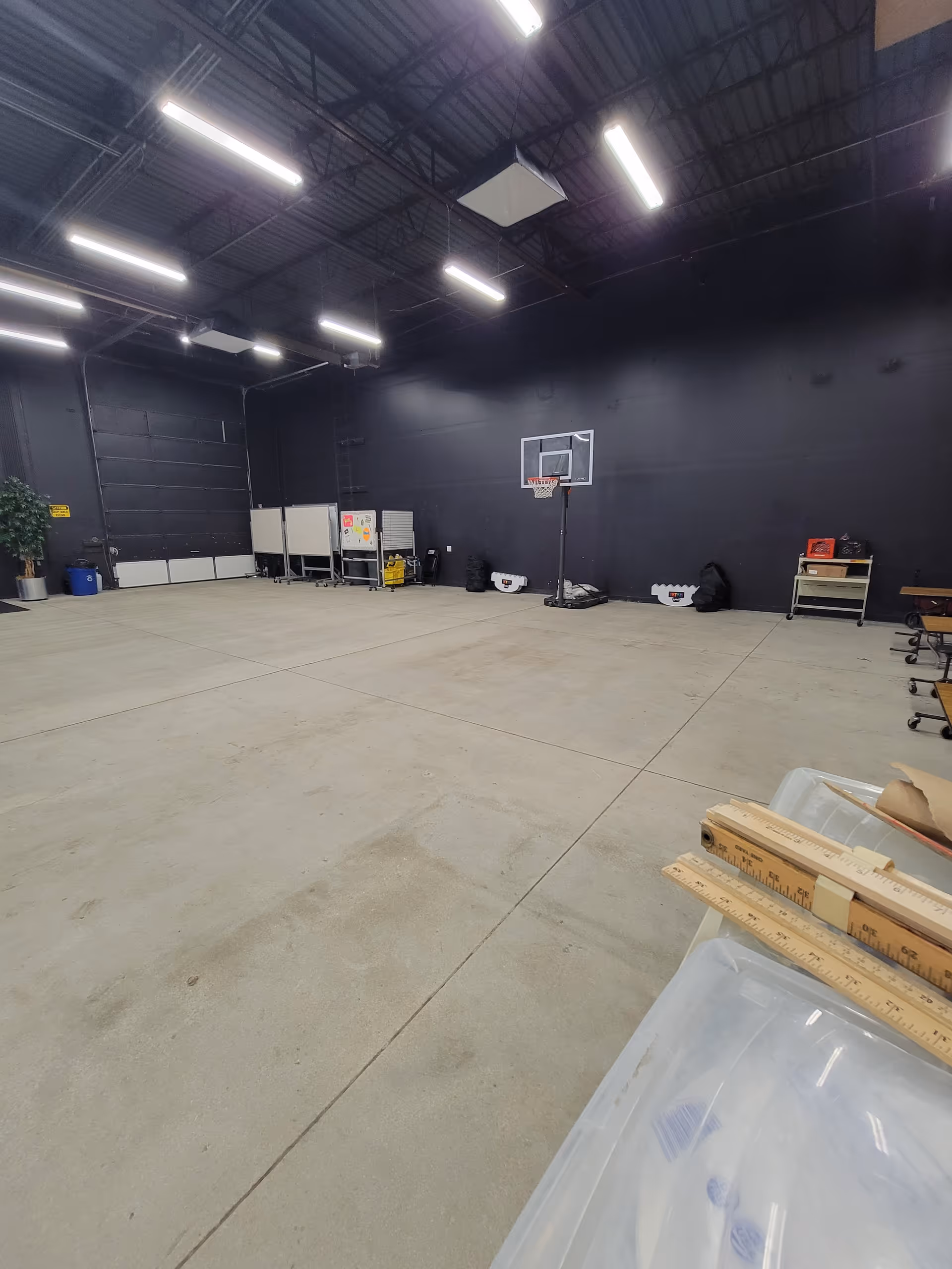 A large empty indoor gym space with a basketball hoop and scattered equipment.