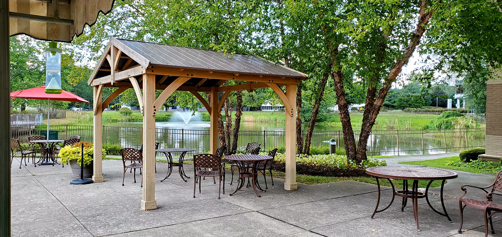 Outdoor patio with metal tables and chairs under a wooden gazebo beside a pond fountain