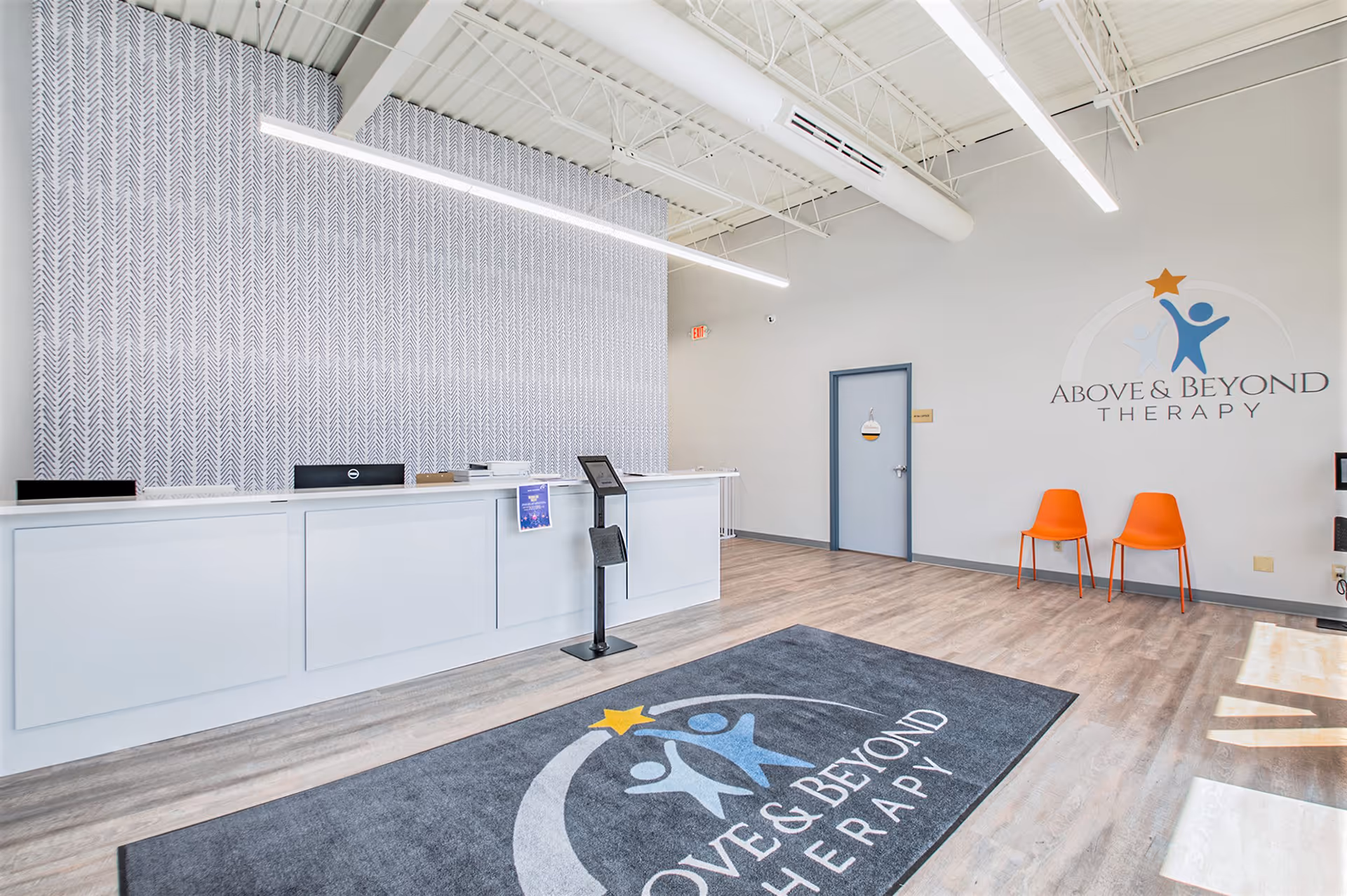 Modern reception area with a long white front desk, logo wall, and orange chairs.