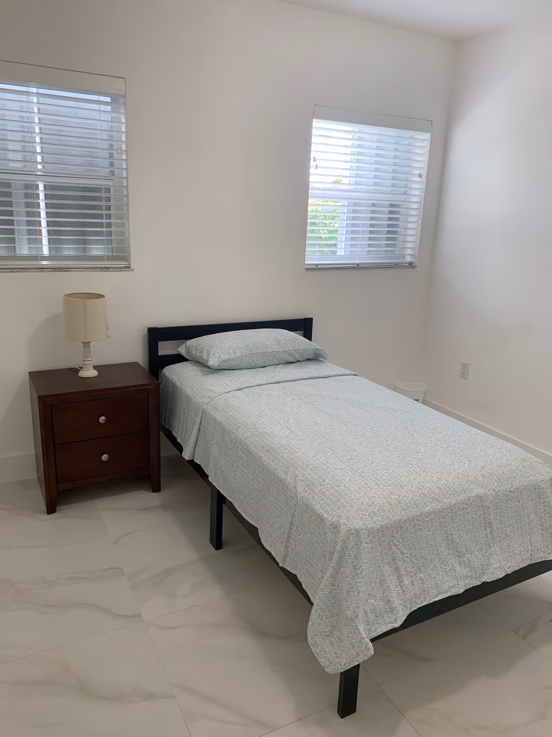 A small bedroom in Karelis Home Care with a single bed, nightstand, lamp, and two windows.