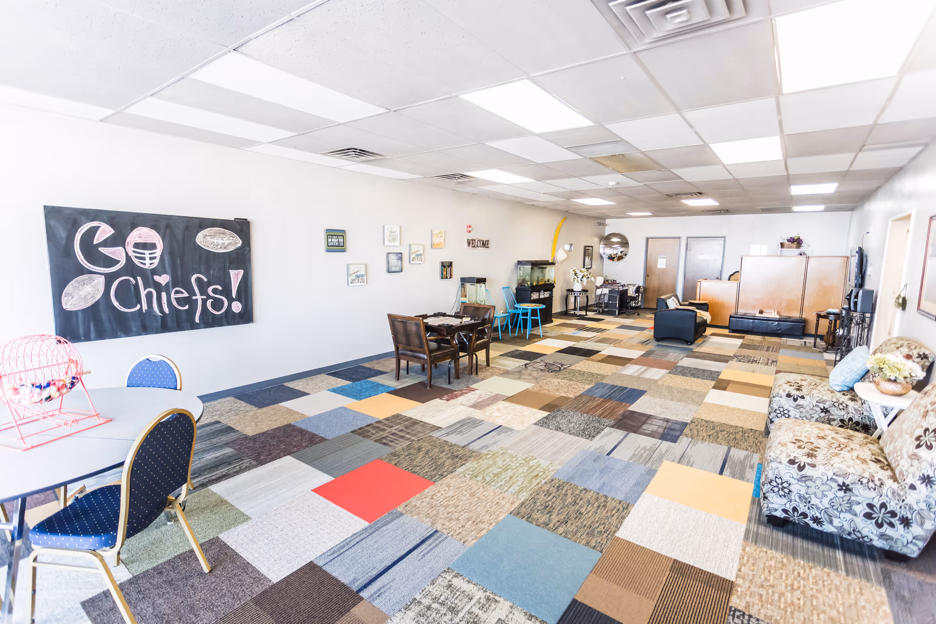 Bright, multi-colored carpeted activity room with chairs, sofas, tables, and a chalkboard reading Go Chiefs.