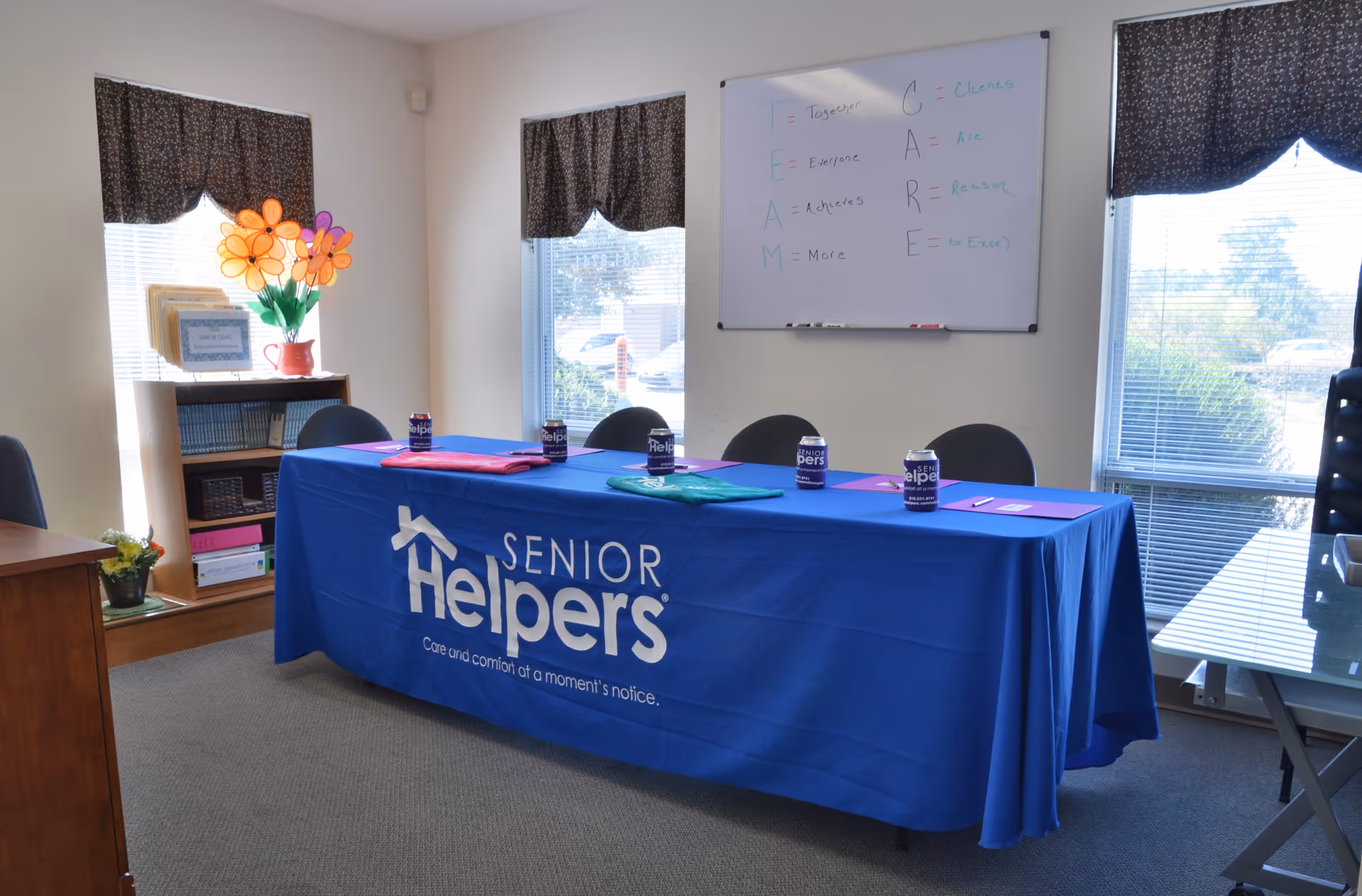 A blue Senior Helpers banner covers a table in a bright, windowed meeting room.