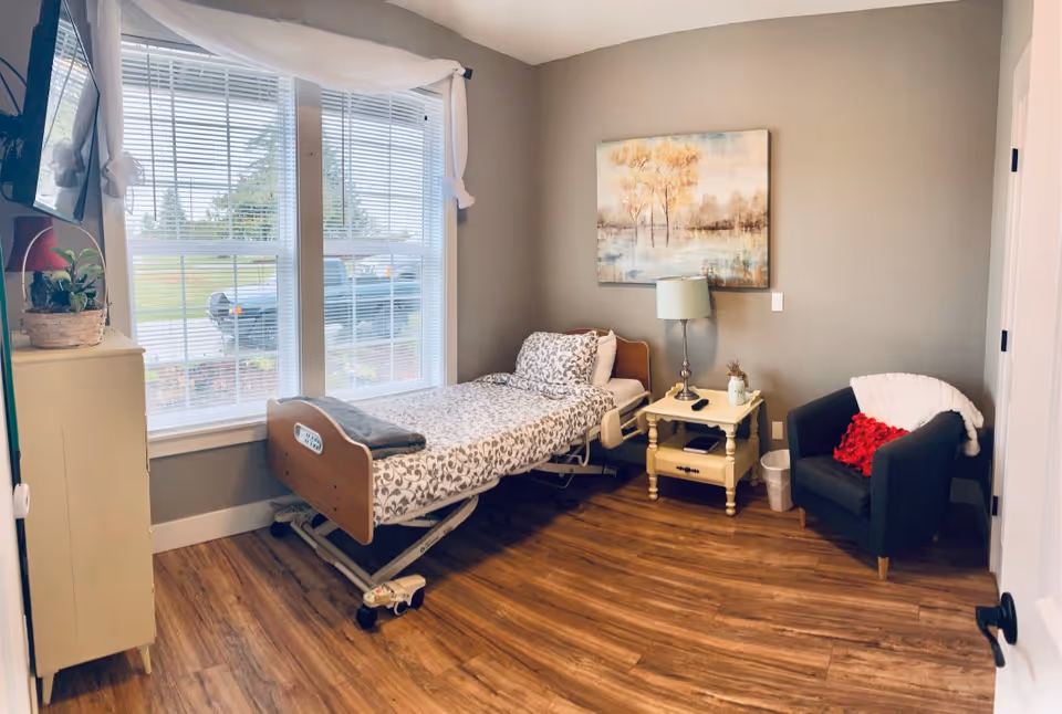 A senior care facility bedroom with an adjustable hospital bed, nightstand, lamp, and chair.