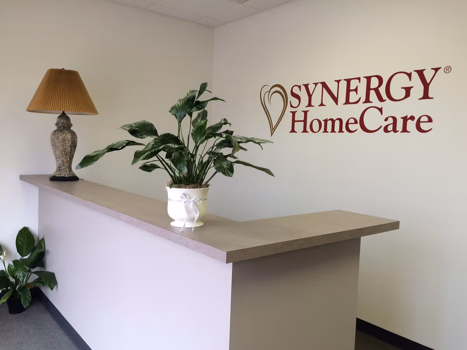 Reception desk with a plant, lamp, and SYNERGY HomeCare logo on the wall.