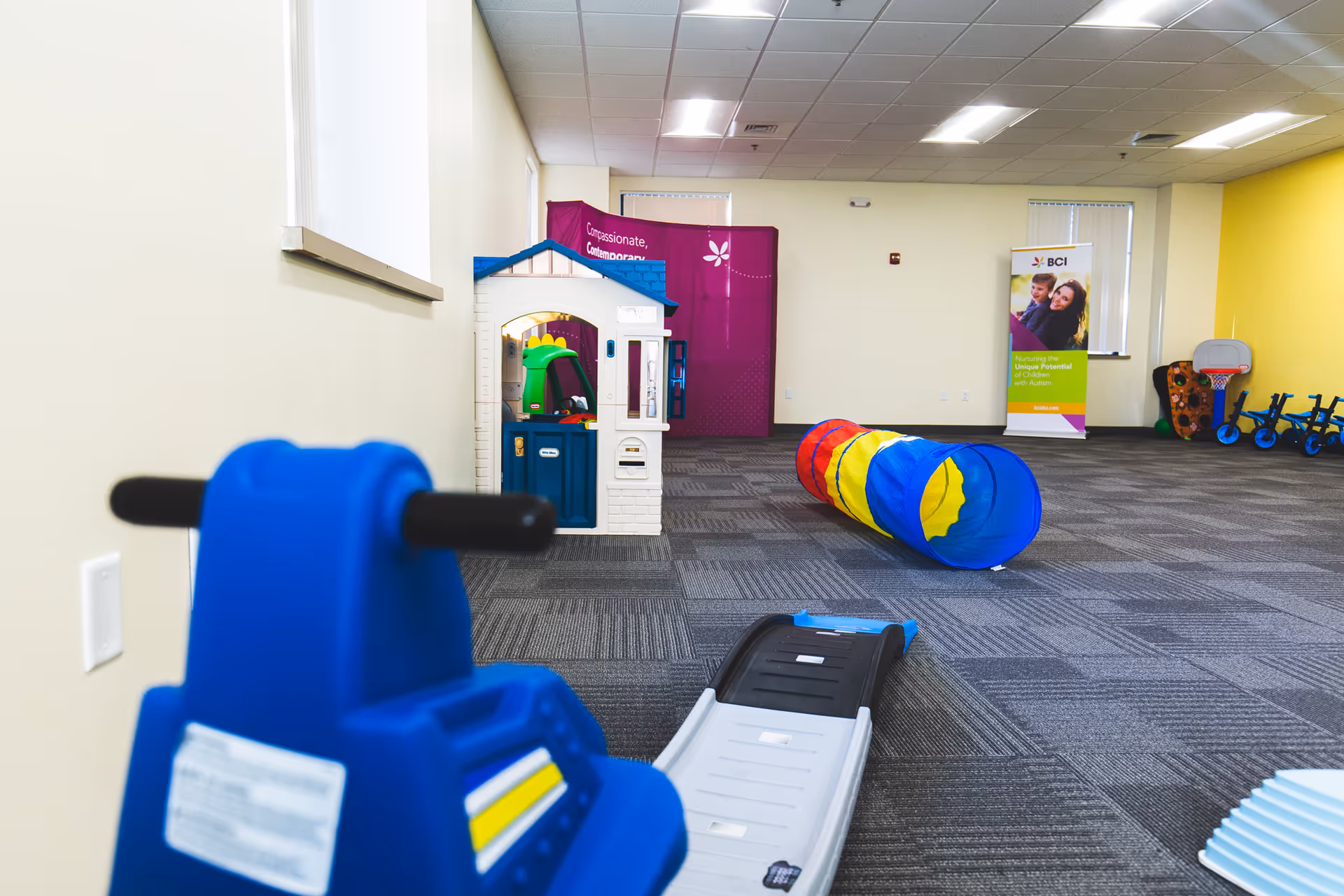 Bright indoor activity room with toy house, tunnel, and play equipment.