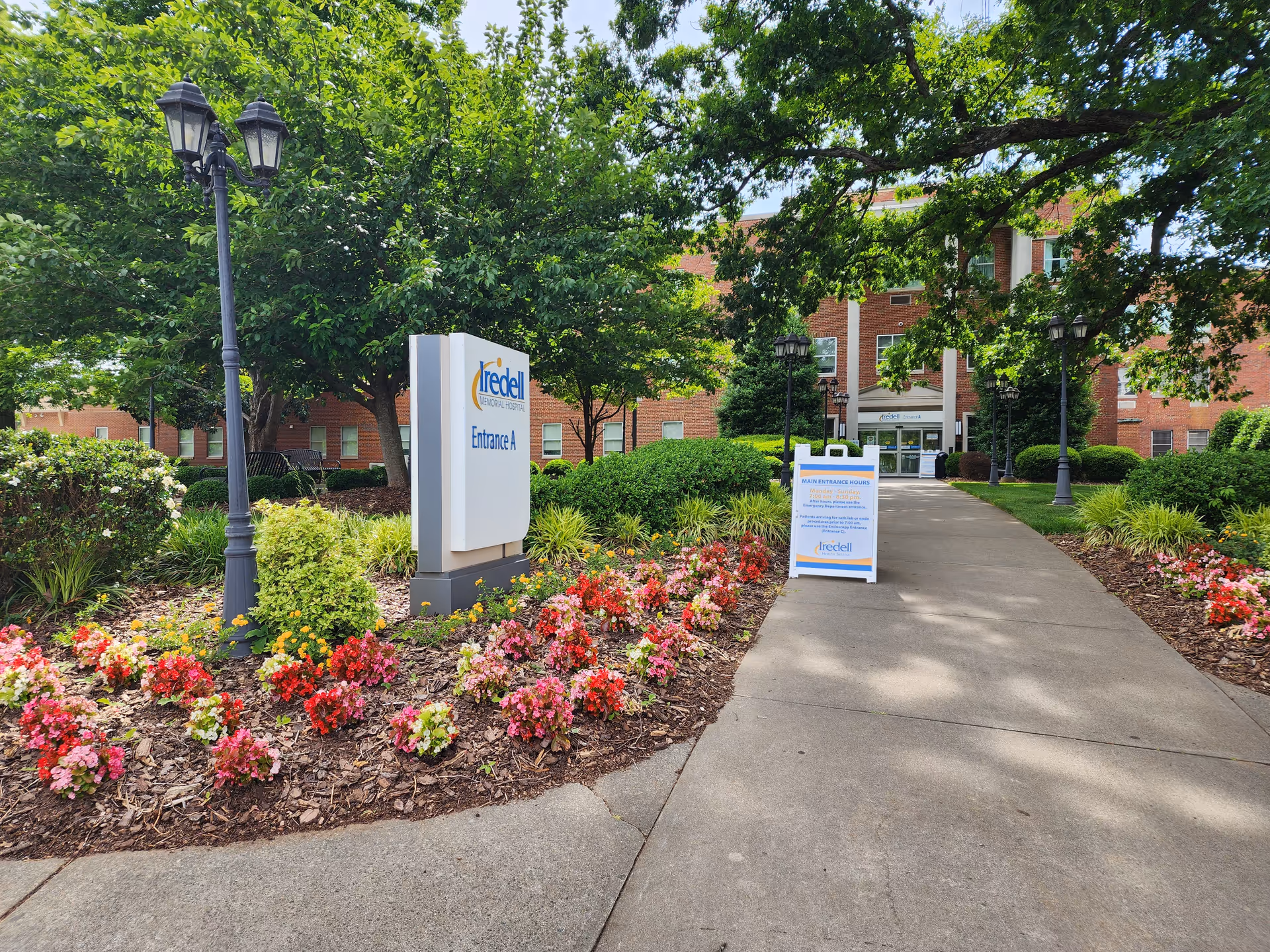 Outdoor walkway to Iredell Health System facility, with flowering beds, trees, and brick building in view.