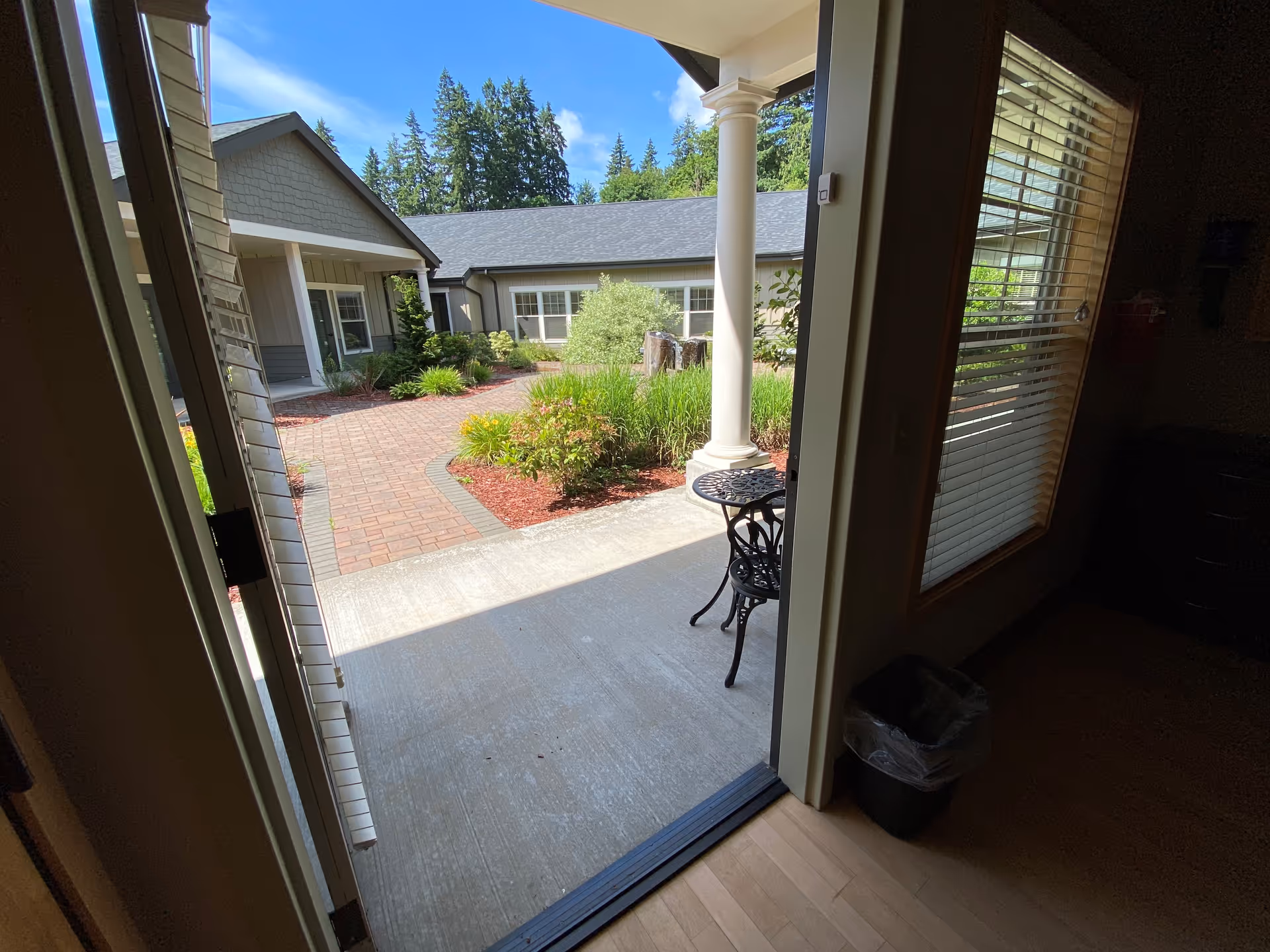 From indoors, a sunny courtyard with brick walkway, shrubs, and outdoor seating.