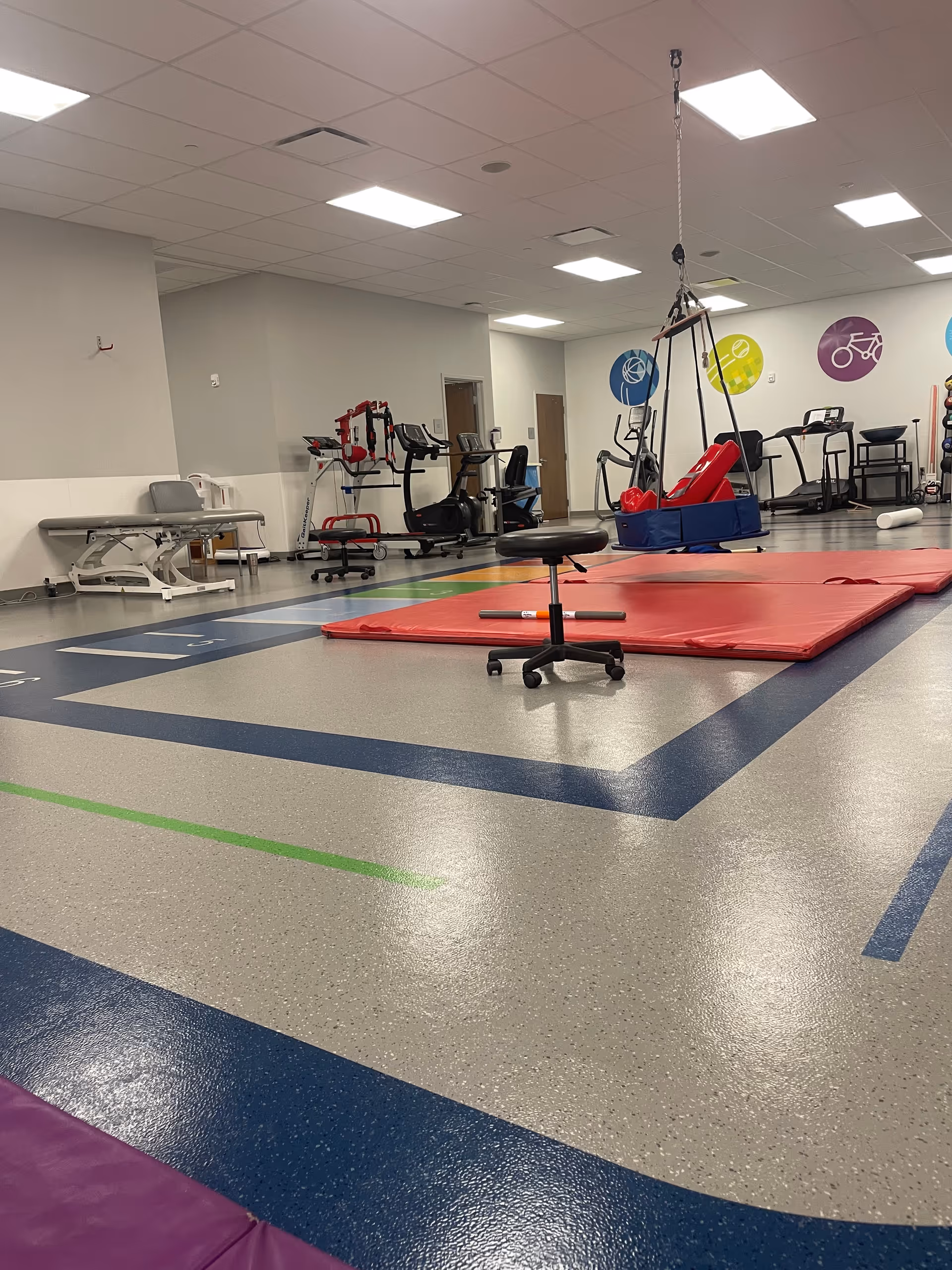 Bright indoor rehabilitation gym with red mats, exercise machines, and suspended therapy apparatus.