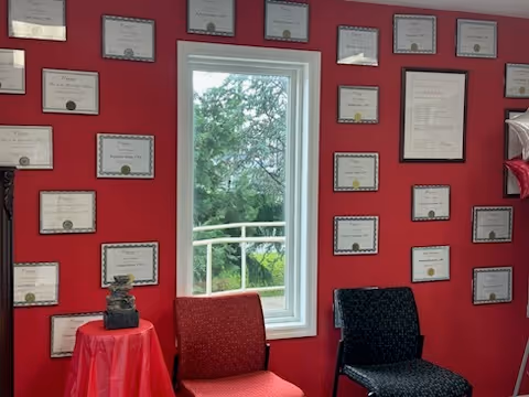 Red wall with many framed certificates around a window and two chairs.