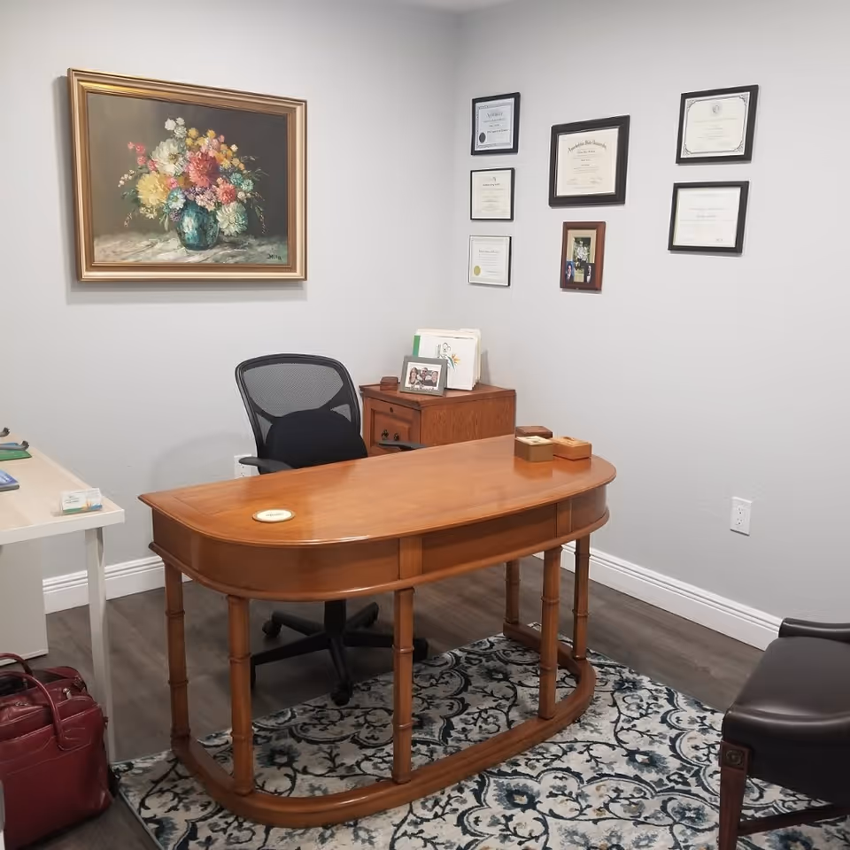 A wooden curved desk with chair in an office, framed certificates on gray walls.