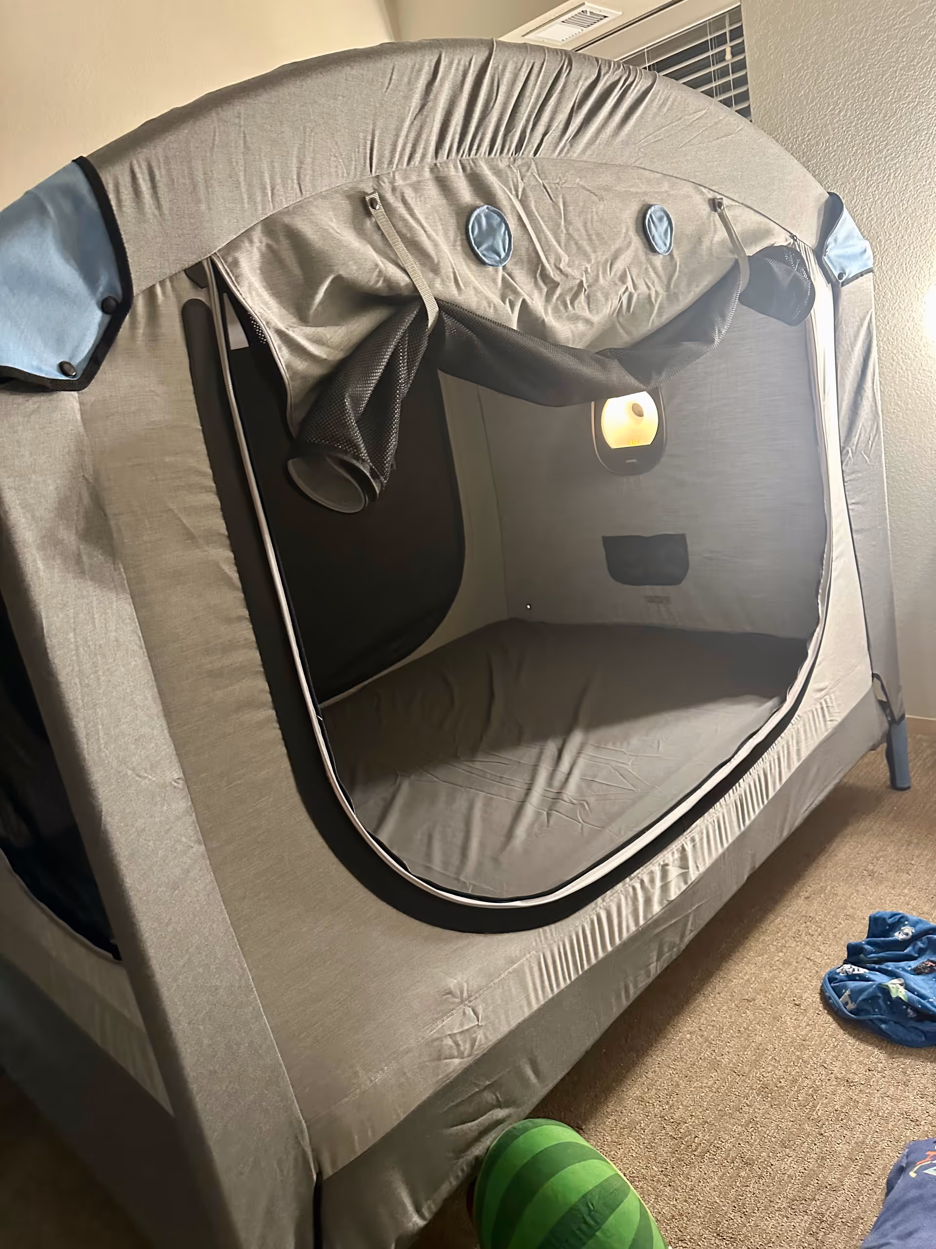 Grey pop-up tent inside a carpeted room with a ball and scattered clothing items.