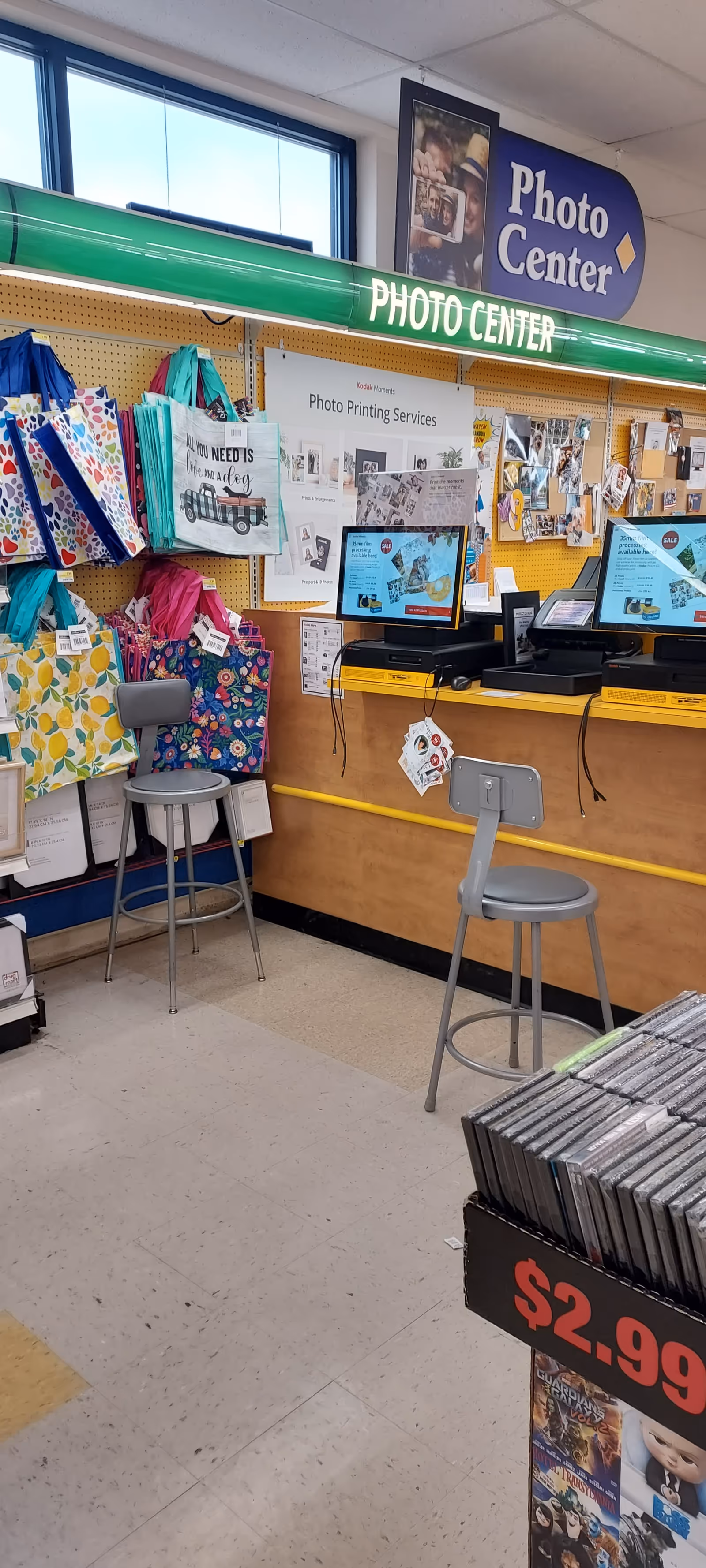 Photo center inside Discount Drug Mart with computers, stools, and colorful tote bags on display.