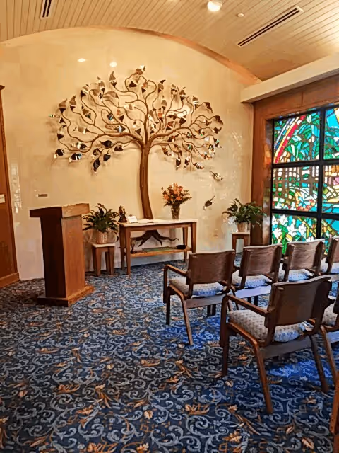 Chapel-like interior with podium, stained-glass window, a tree wall sculpture, and arranged chairs.