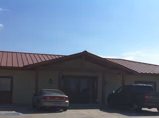 Single-story building with a red metal roof and two parked cars at the entrance.