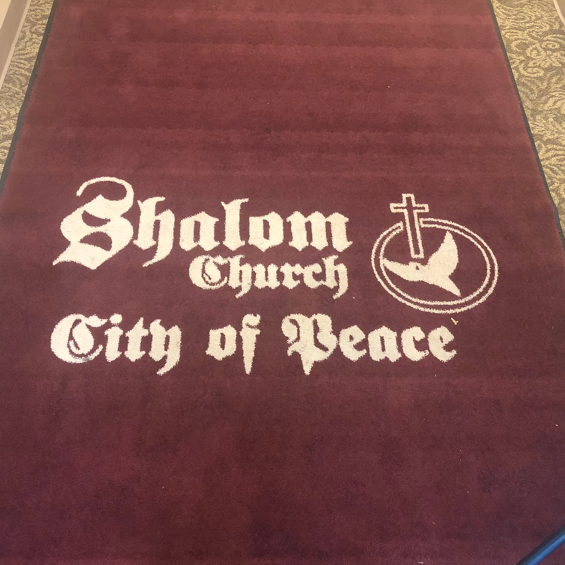 Maroon hallway carpet featuring Shalom Church City of Peace logo and cross.