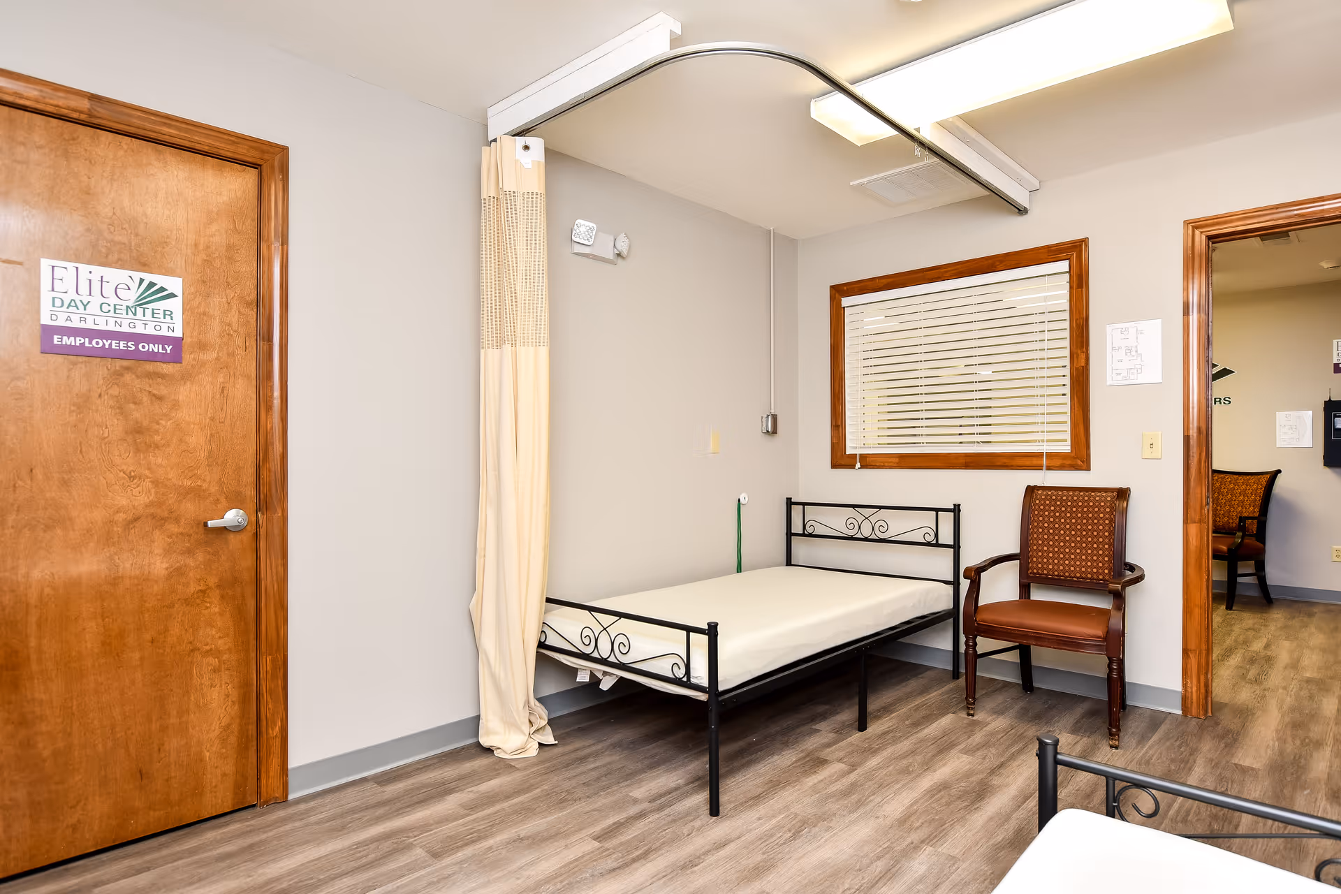 A single bed with metal frame, privacy curtain, window, and wooden chair in a resident room.