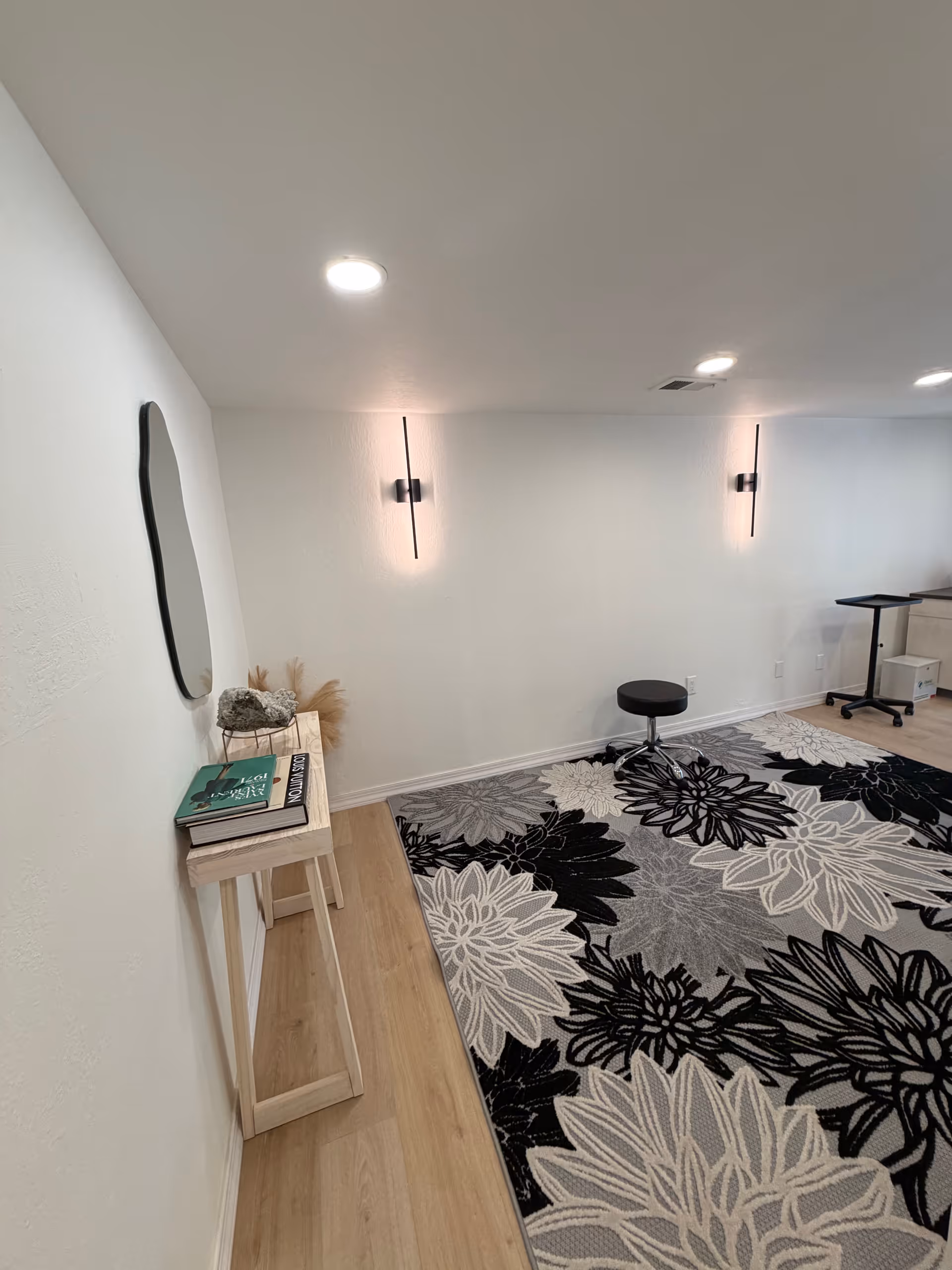 A narrow interior hallway with white walls, wall sconces, and a floral rug.
