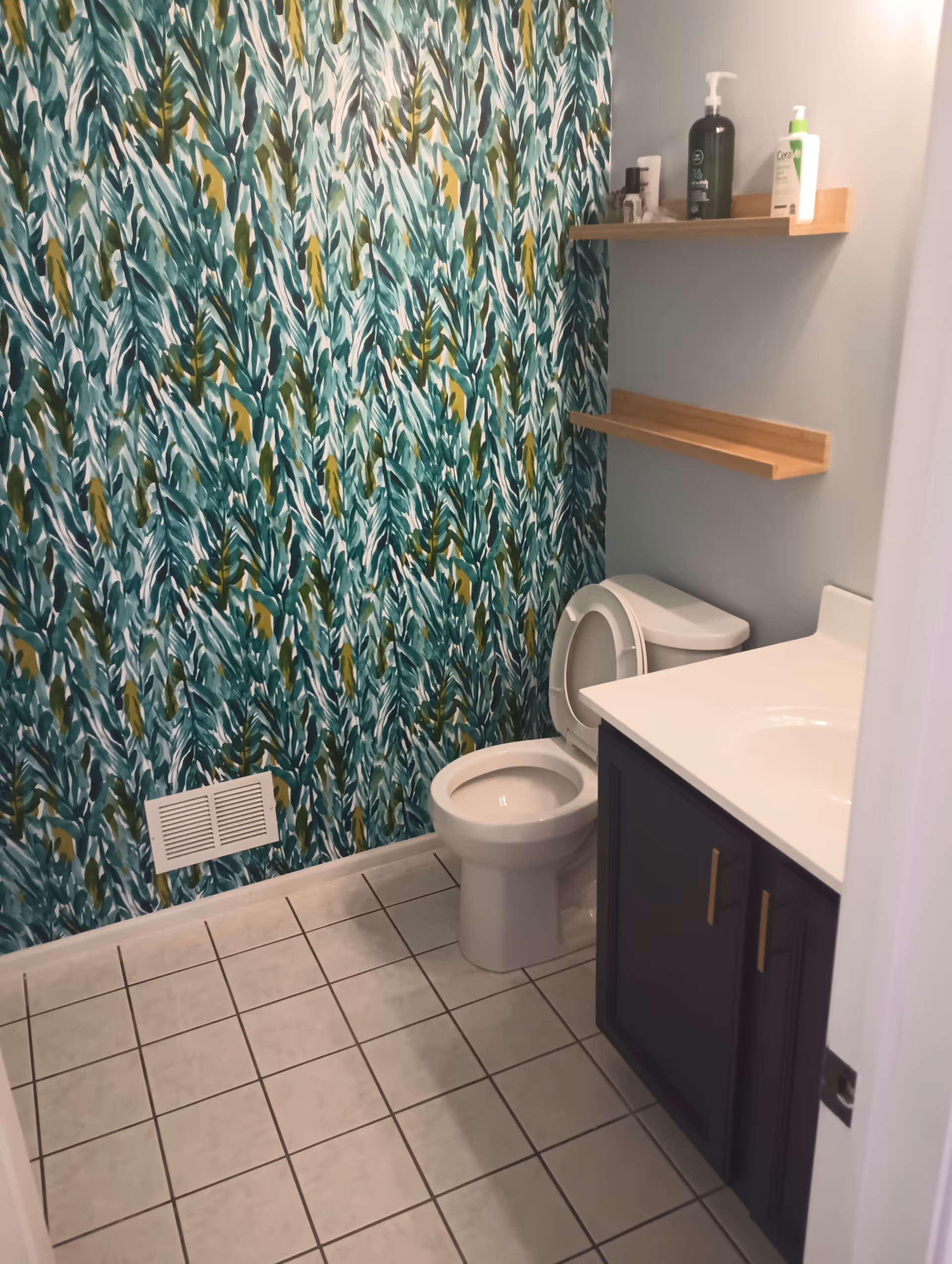 Small bathroom with leafy teal wallpaper, toilet, sink, and two wooden shelves.
