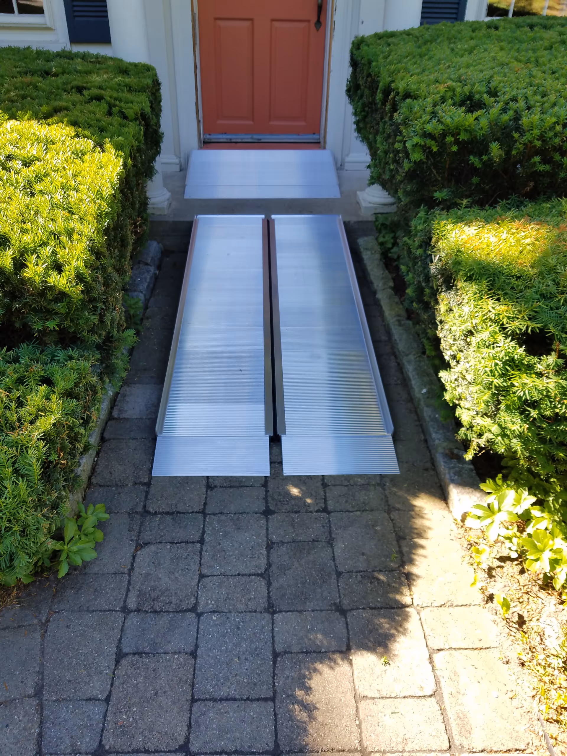 Exterior aluminum ramp leading to a red front door flanked by hedges