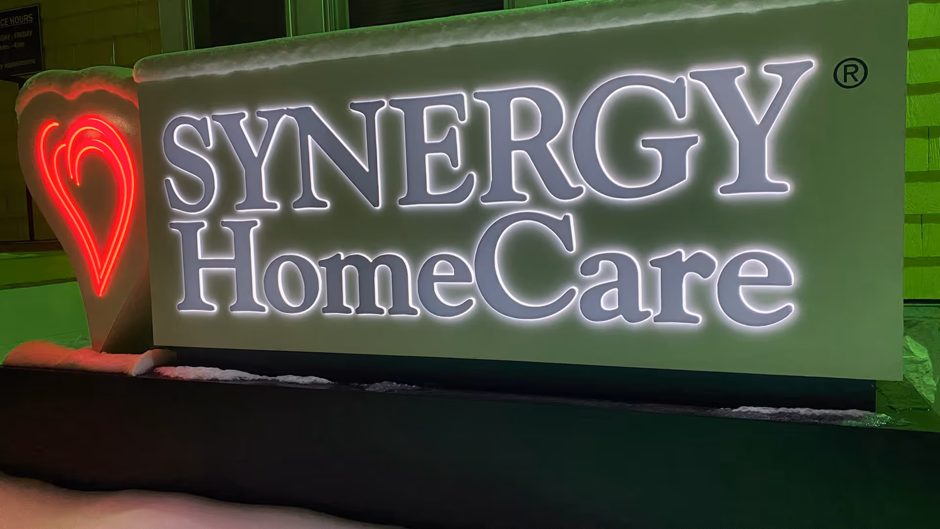 Illuminated SYNERGY HomeCare sign with red heart logo on a building front.