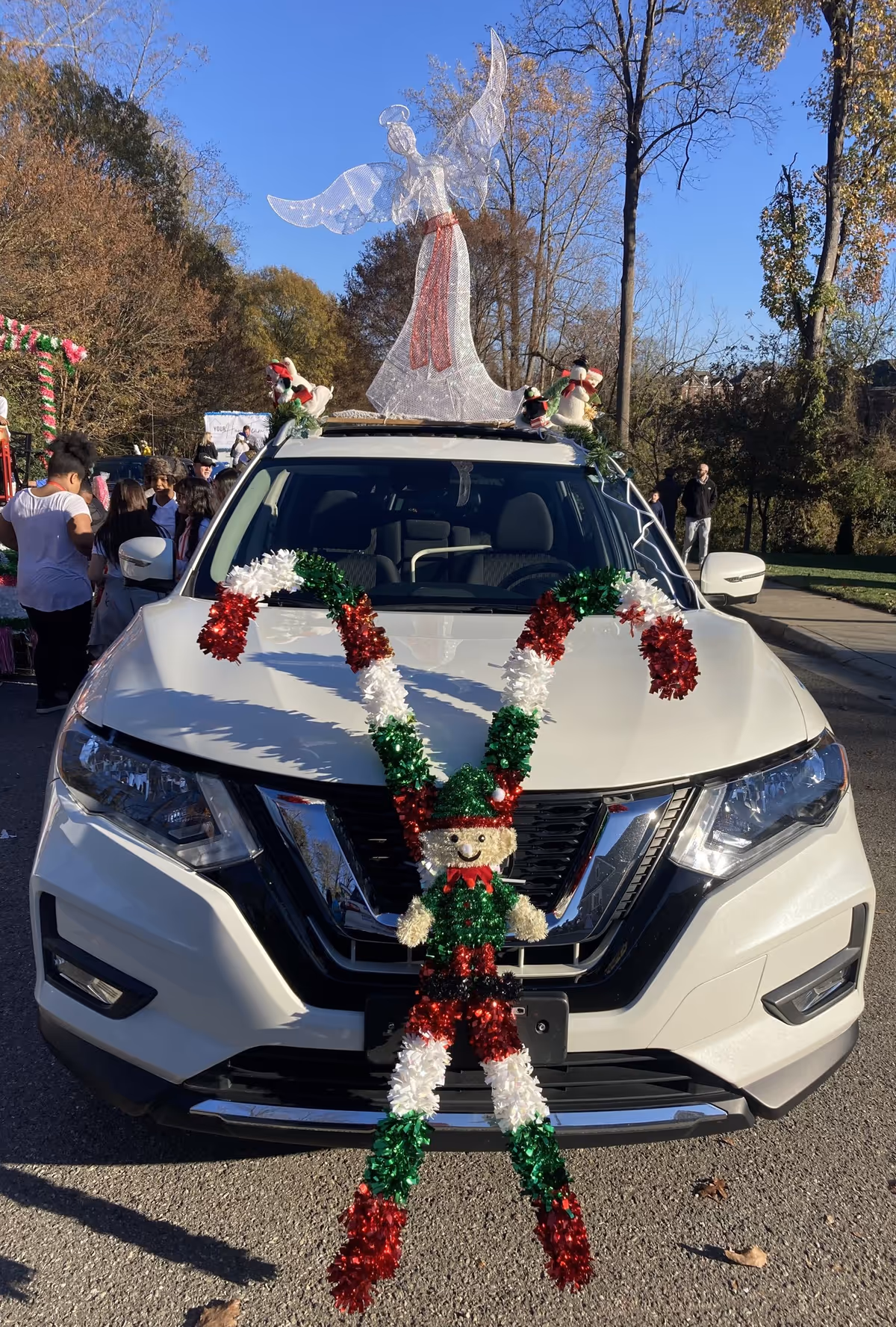Decorated white SUV with garlands and an angel figure on the roof at a parade.