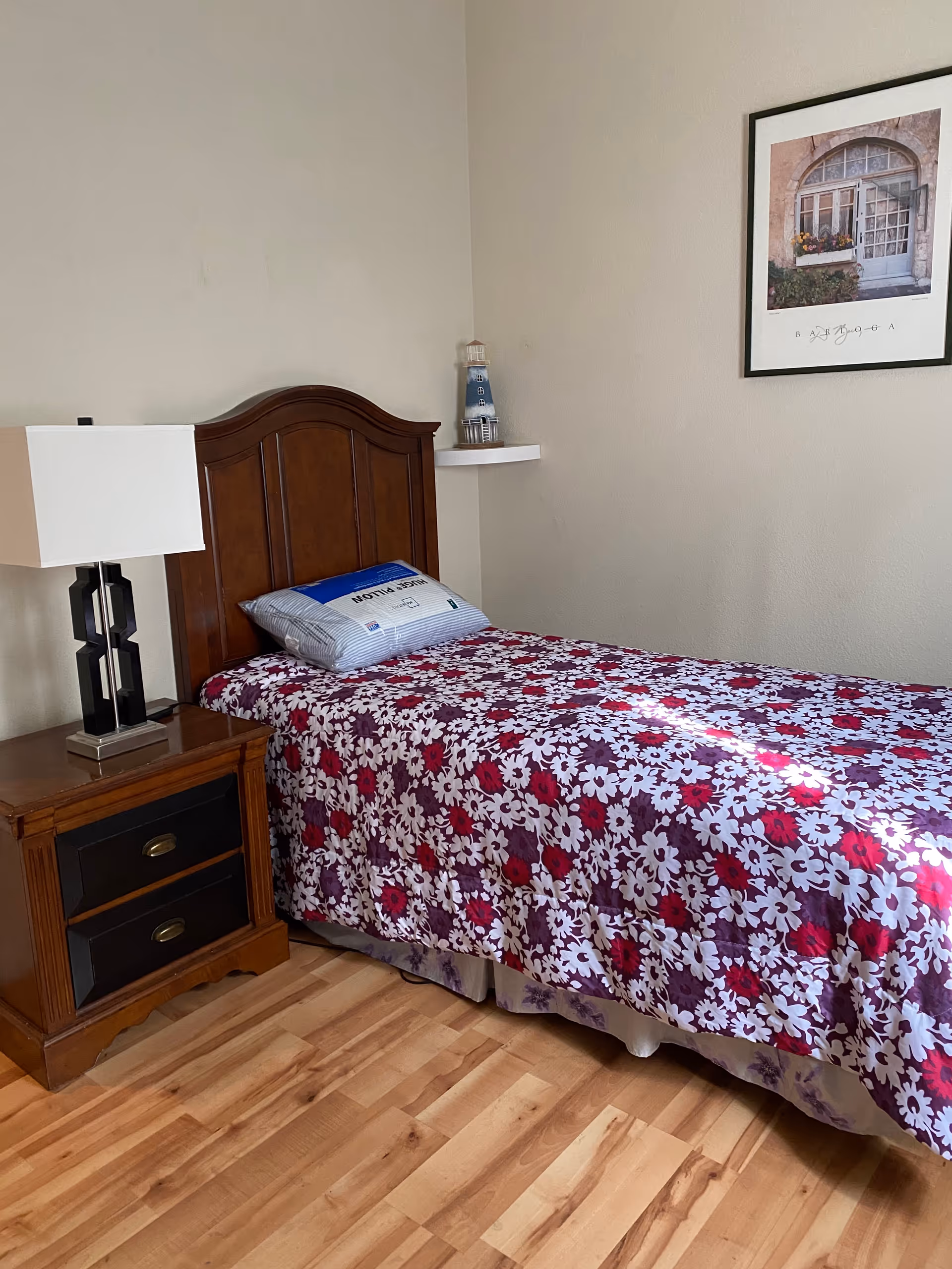Single bedroom with wooden headboard, floral quilt, nightstand, lamp, and wall art.