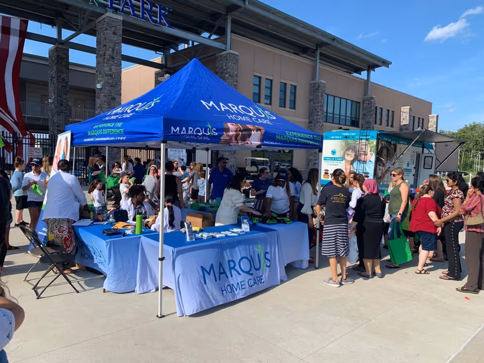 Outdoor event with Marquis Home Care tent and crowd gathered around informational tables.