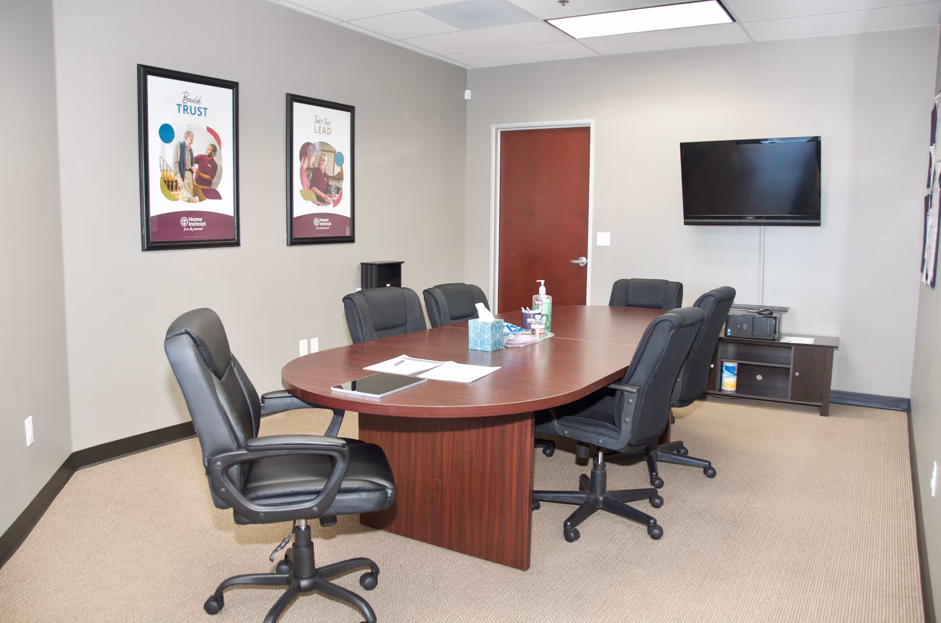 An office-style conference room with a large curved table, black chairs, and a wall-mounted TV.