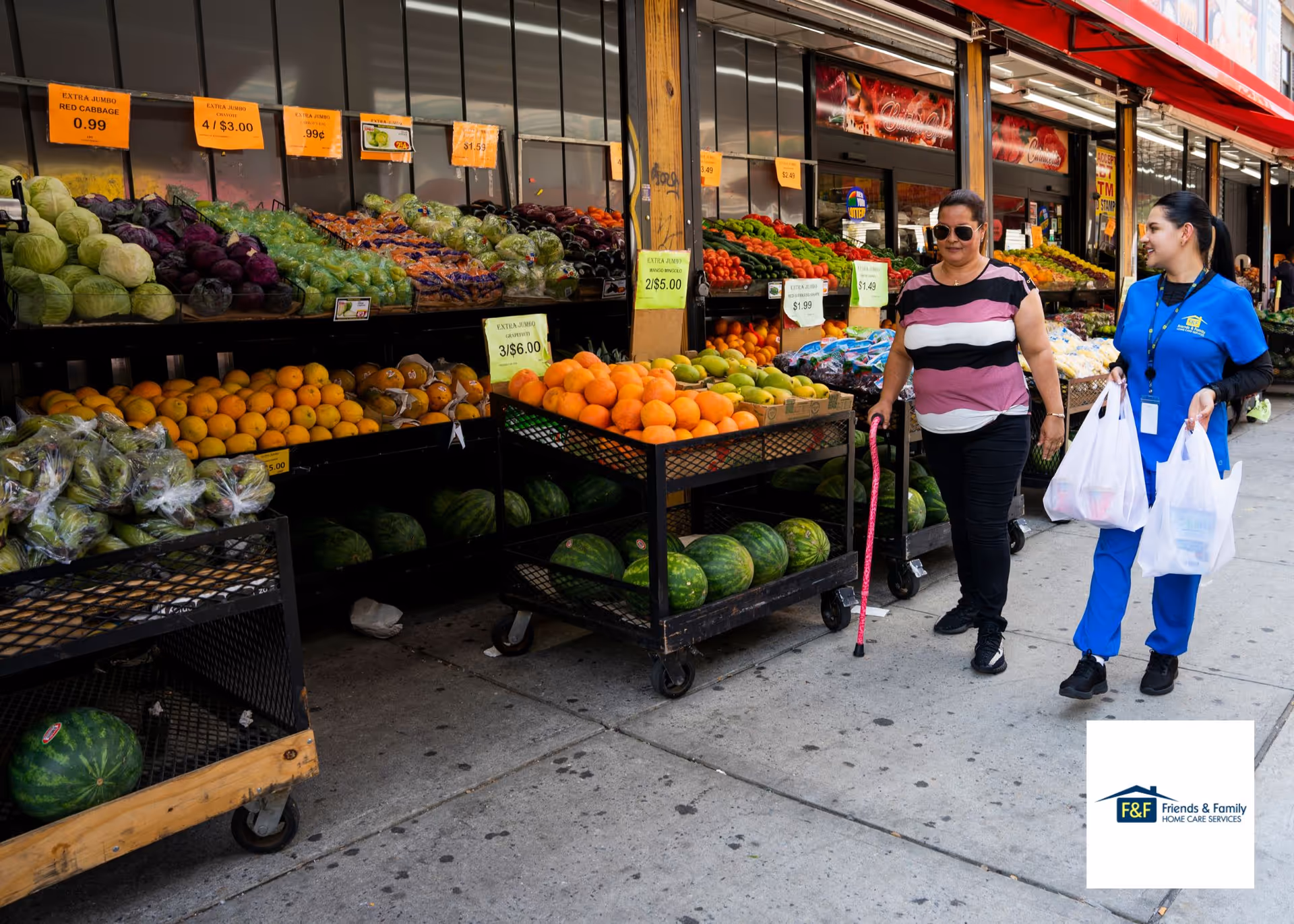 Two women walk past a colorful fruit storefront with produce on display outside.