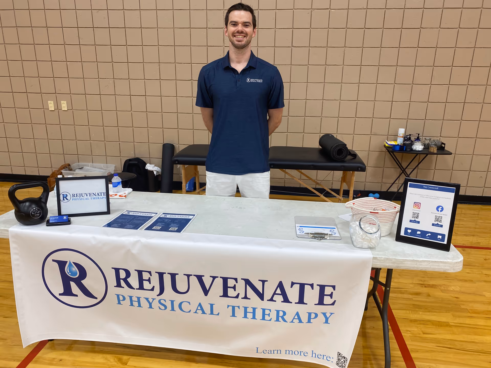 A staff member stands behind a table with a Rejuvenate Physical Therapy banner.