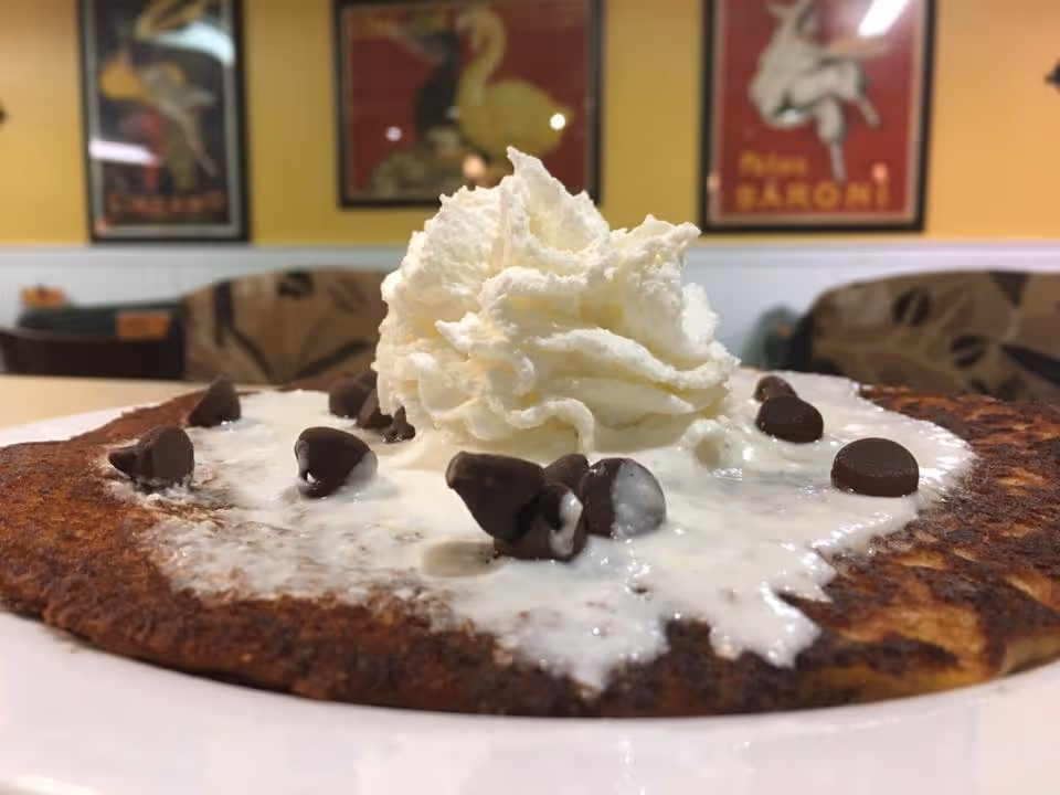 Close up of a chocolate chip pancake topped with whipped cream in a dining area.