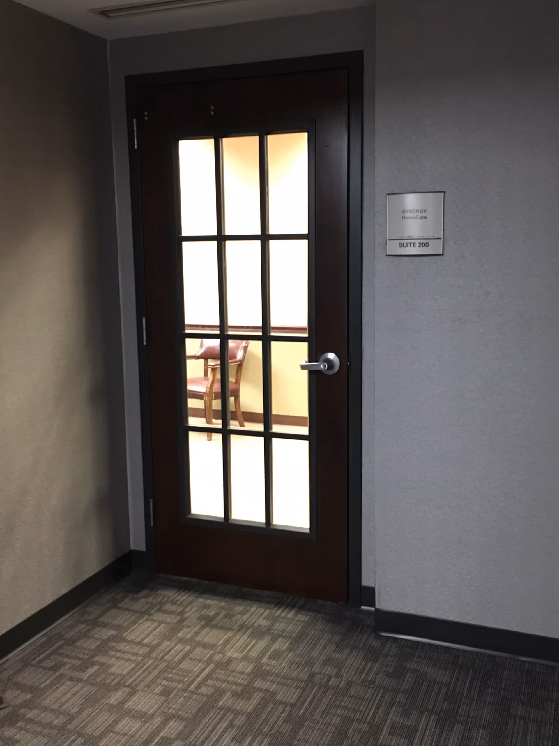 Interior hallway with a wooden glass-paneled door to Synergy HomeCare Suite 200.