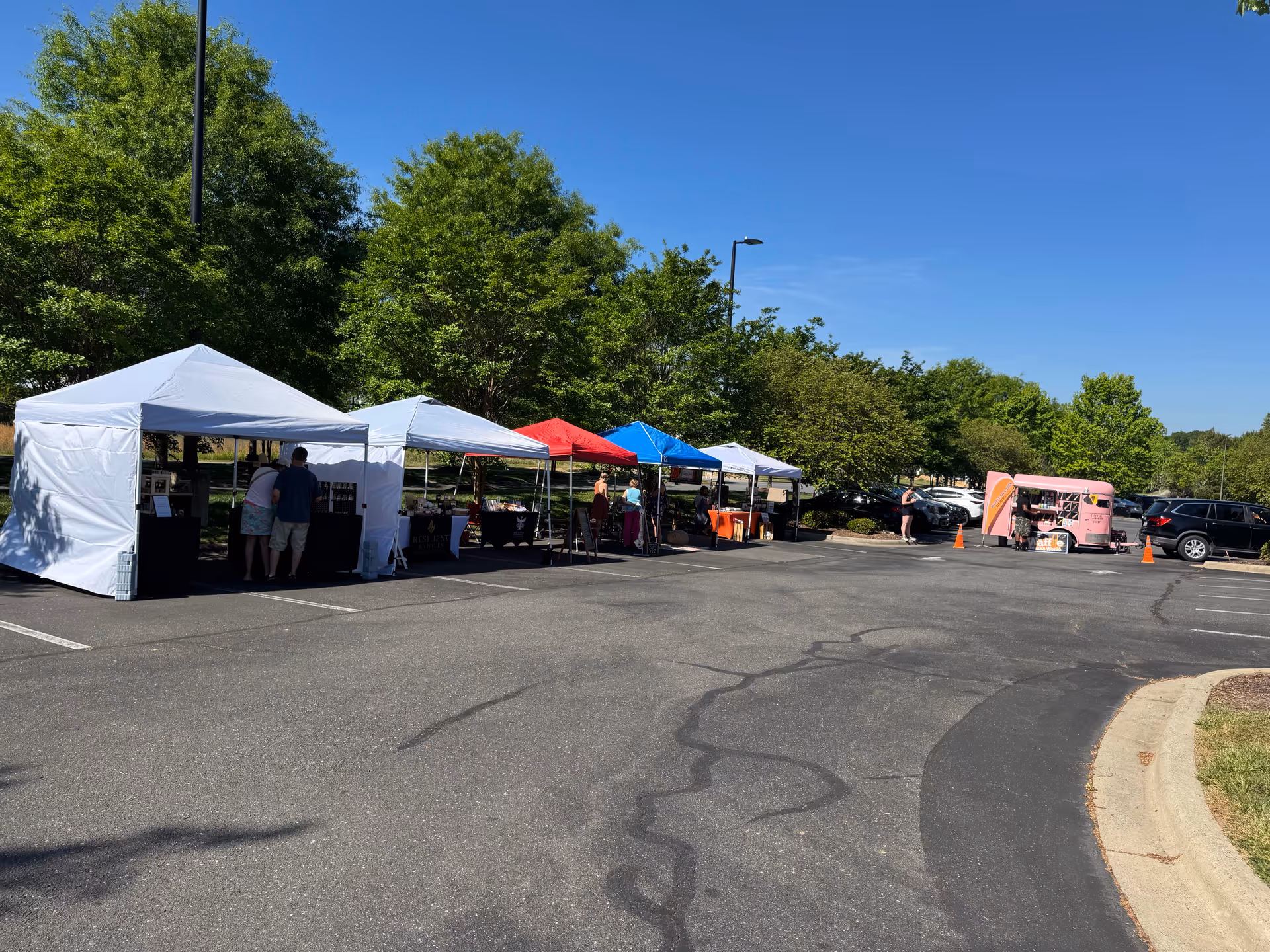 Tents set up in a parking lot beside greenery, with people browsing vendors.