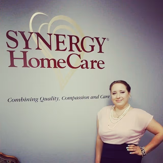 A smiling staff member stands beside a Synergy HomeCare wall logo in a pastel lobby.