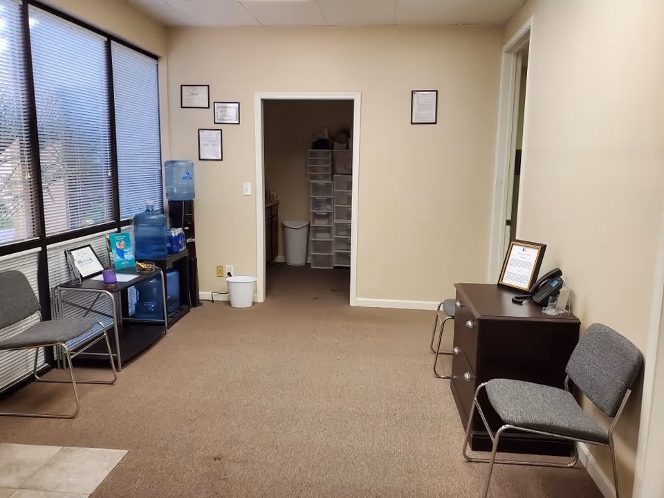Reception area with chairs, water cooler, certificate frames, doorway to storage room, and desk.