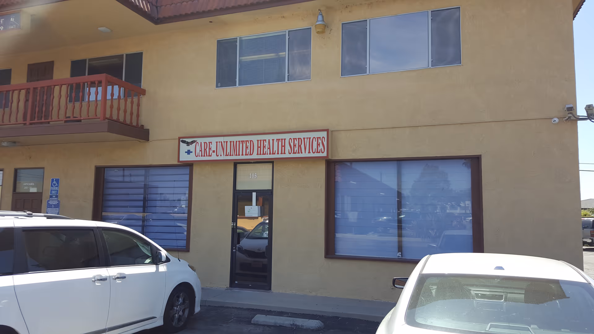 Two-story beige building with a red balcony, sign reading Care Unlimited Health Services, cars parked.