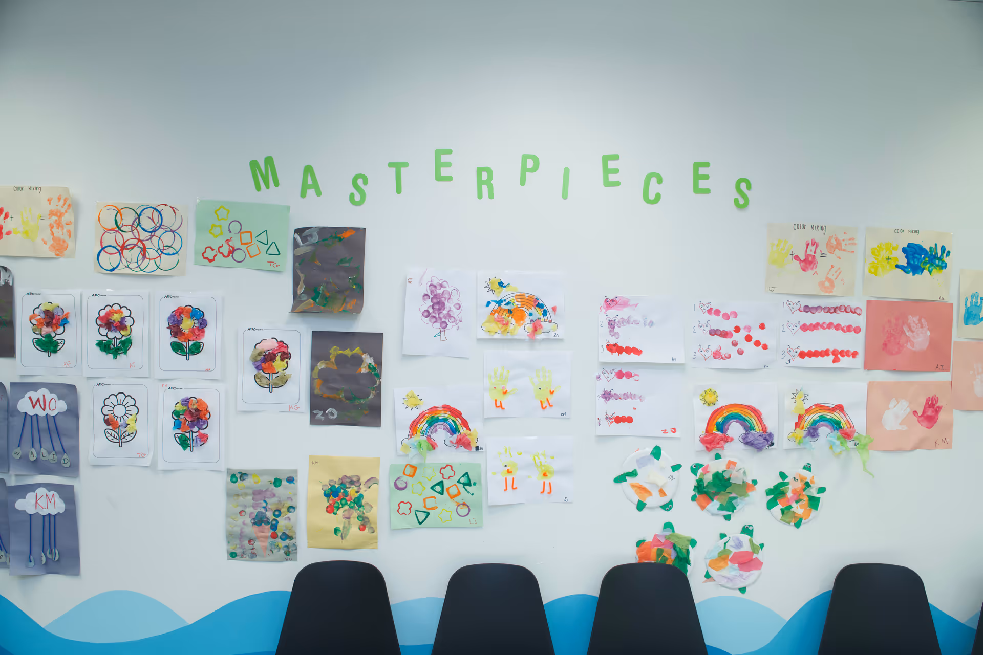 White wall gallery of children's art with 'MASTERPIECES' banner and dark chairs along bottom.