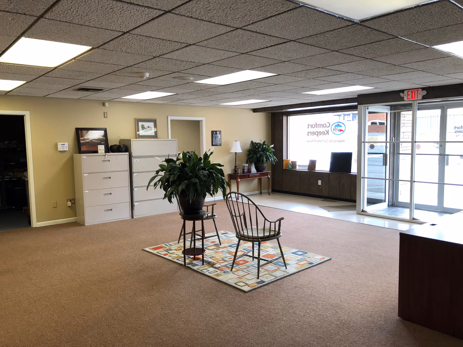 A spacious lobby with chairs, potted plants, filing cabinets, and a reception desk near glass doors.