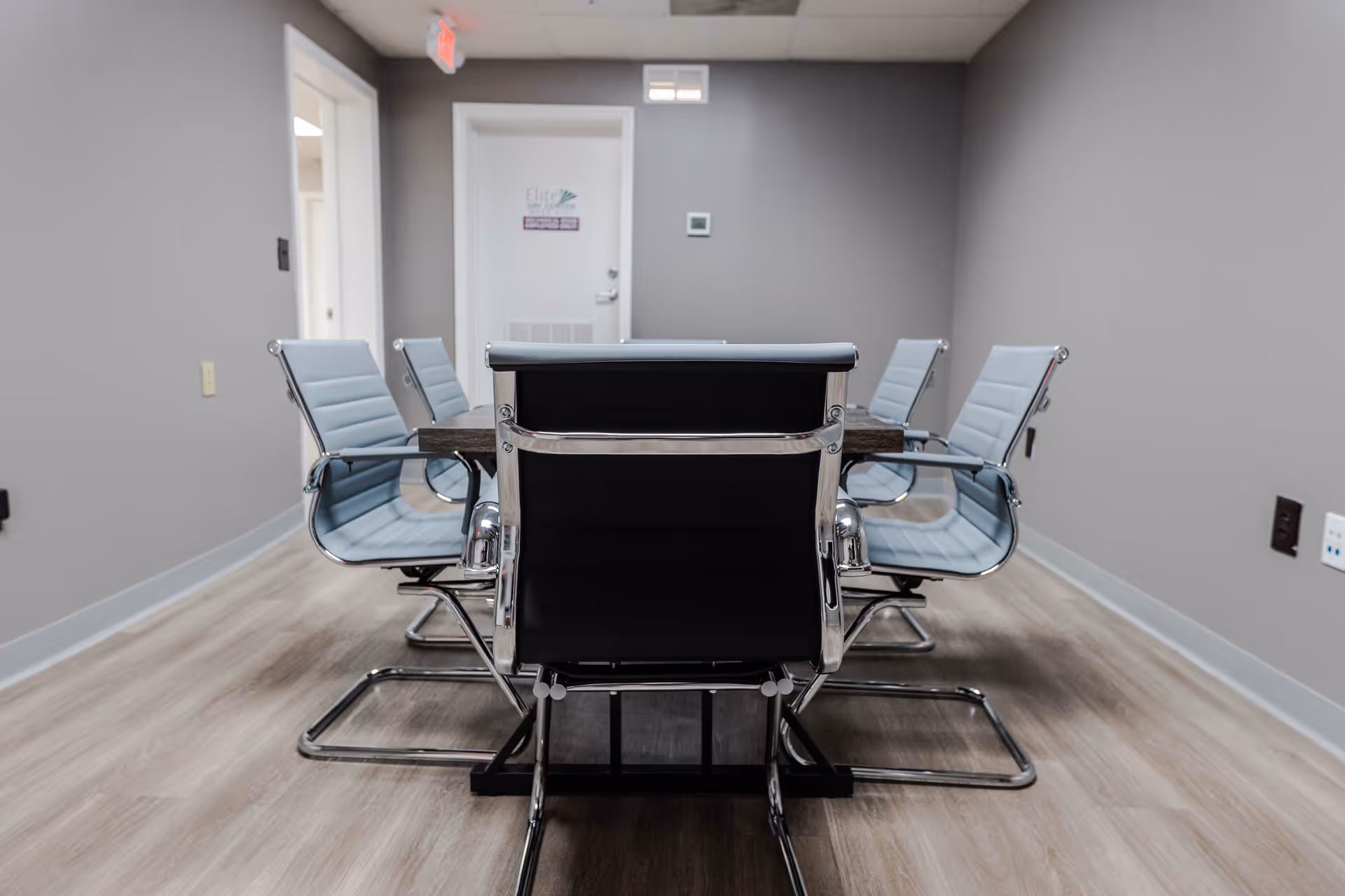 A gray-walled conference room with a dark wooden table and light blue chairs.