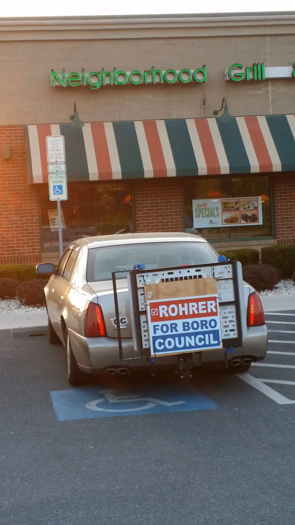 Silver sedan with a campaign sign parked in a handicapped space before striped-awning restaurant.