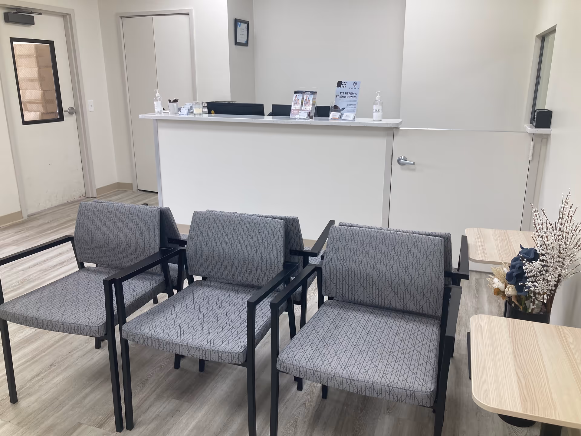 Reception desk behind four gray chairs, small tables, and decorative plant in a clean waiting area.