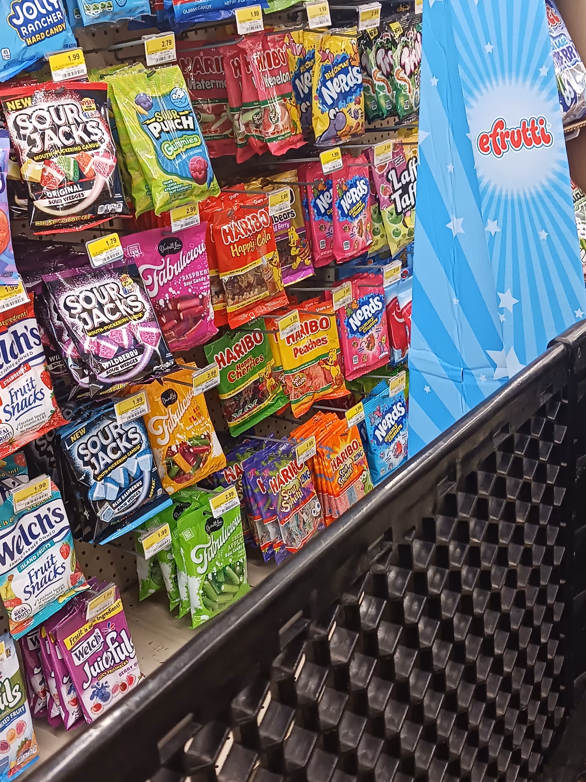 Colorful candy bags line shelves in a Discount Drug Mart aisle beside a black shopping cart.