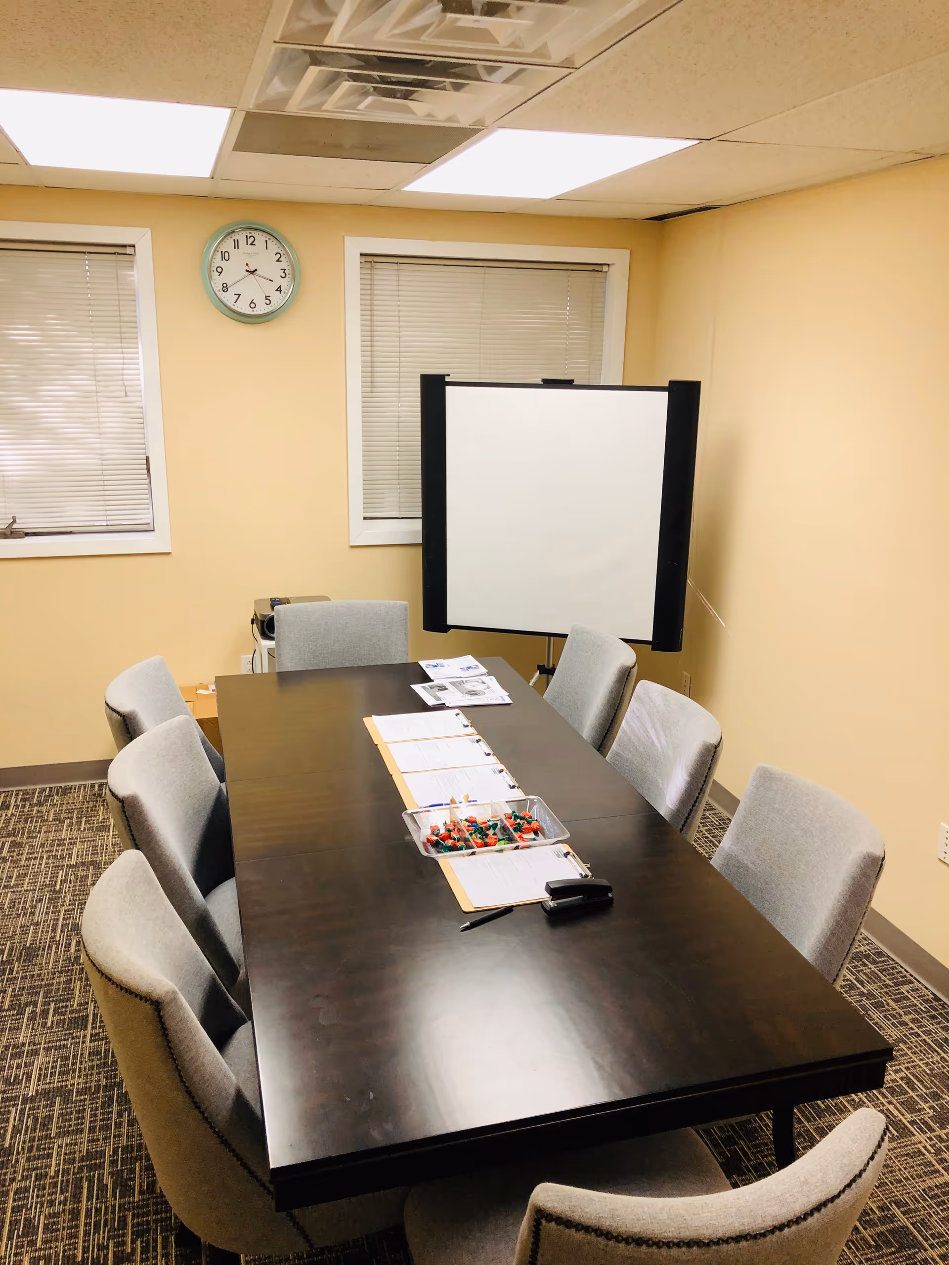 Conference room with long dark table, gray chairs, whiteboard, clock on beige walls.