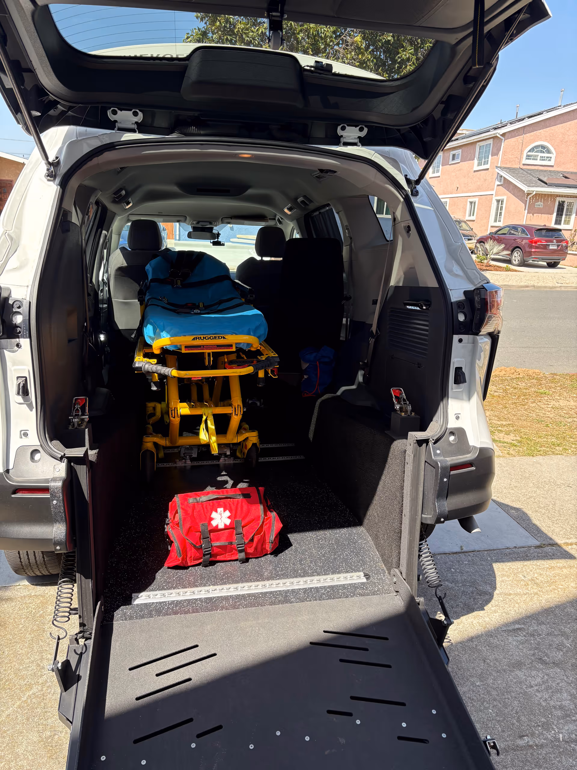 Open vehicle rear showing yellow wheeled stretcher and red medical bag inside.