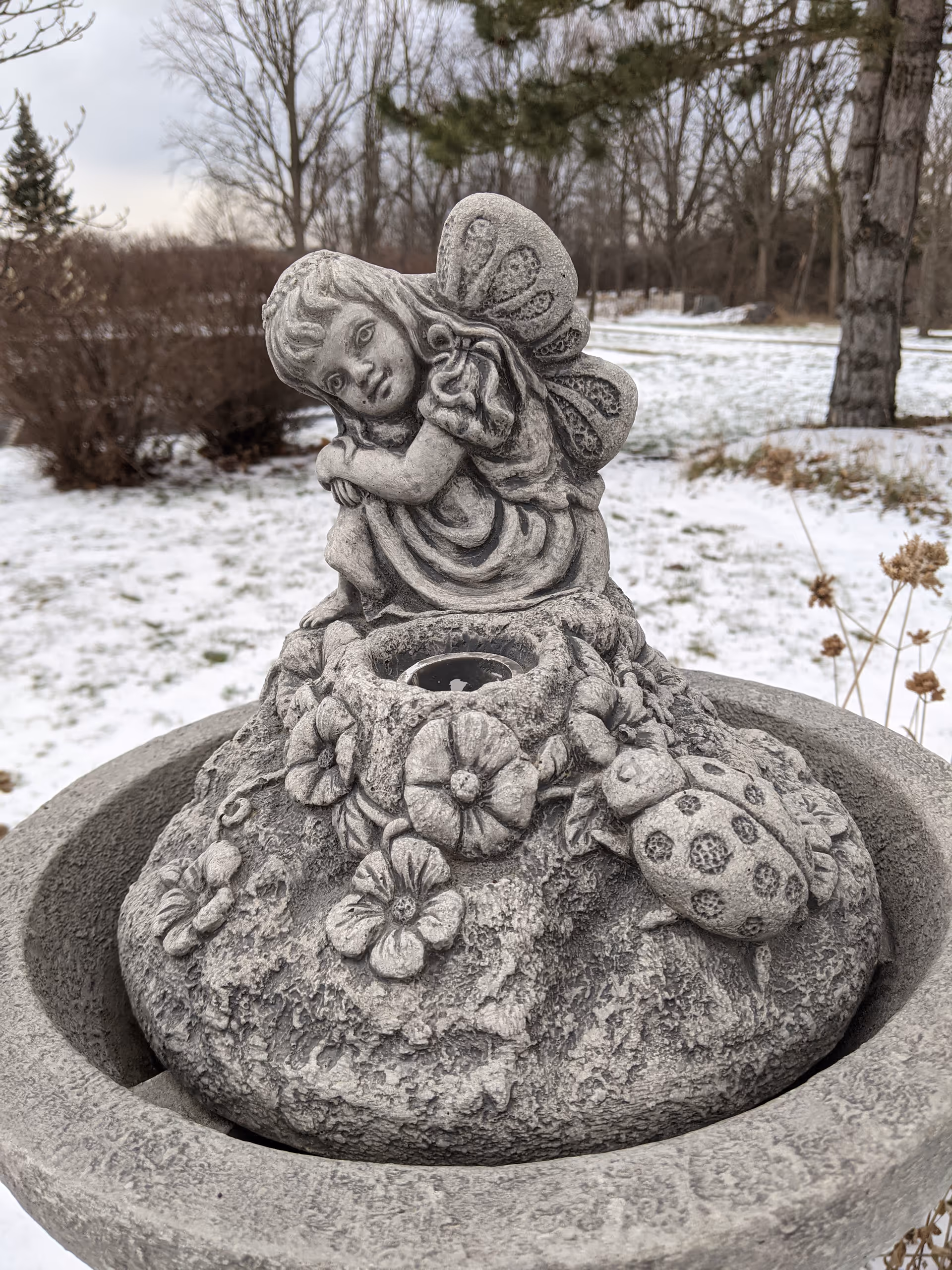 Stone angel statue perched on a fountain in a snowy outdoor garden.