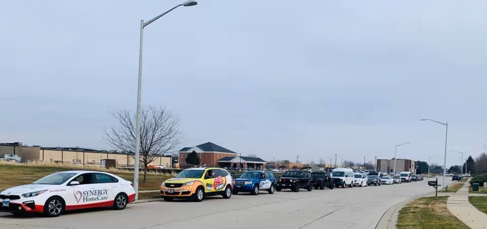 A line of SYNERGY HomeCare vehicles parked along a street in front of a facility.