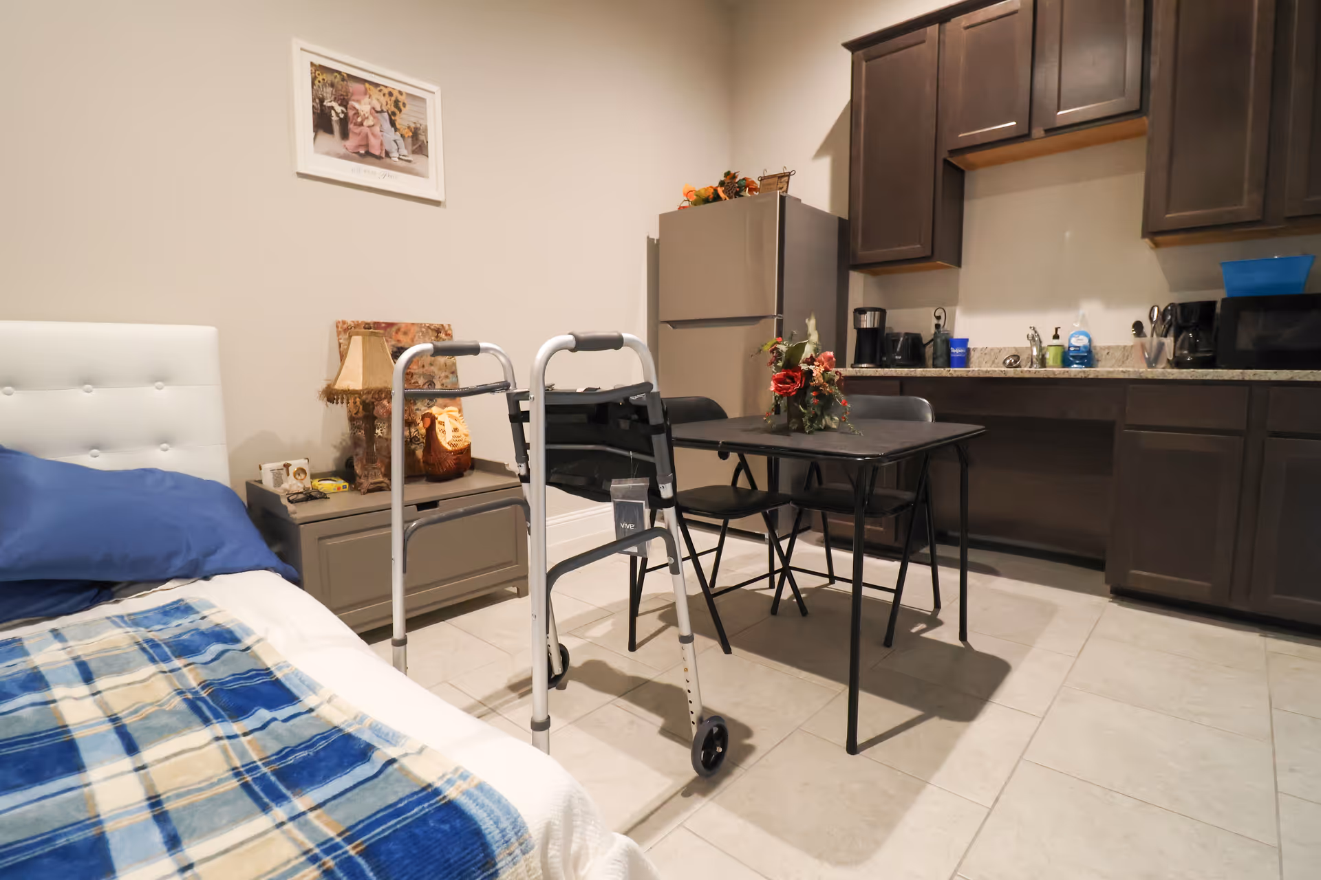 Interior of a senior living studio with bed, walker, table, and kitchenette.